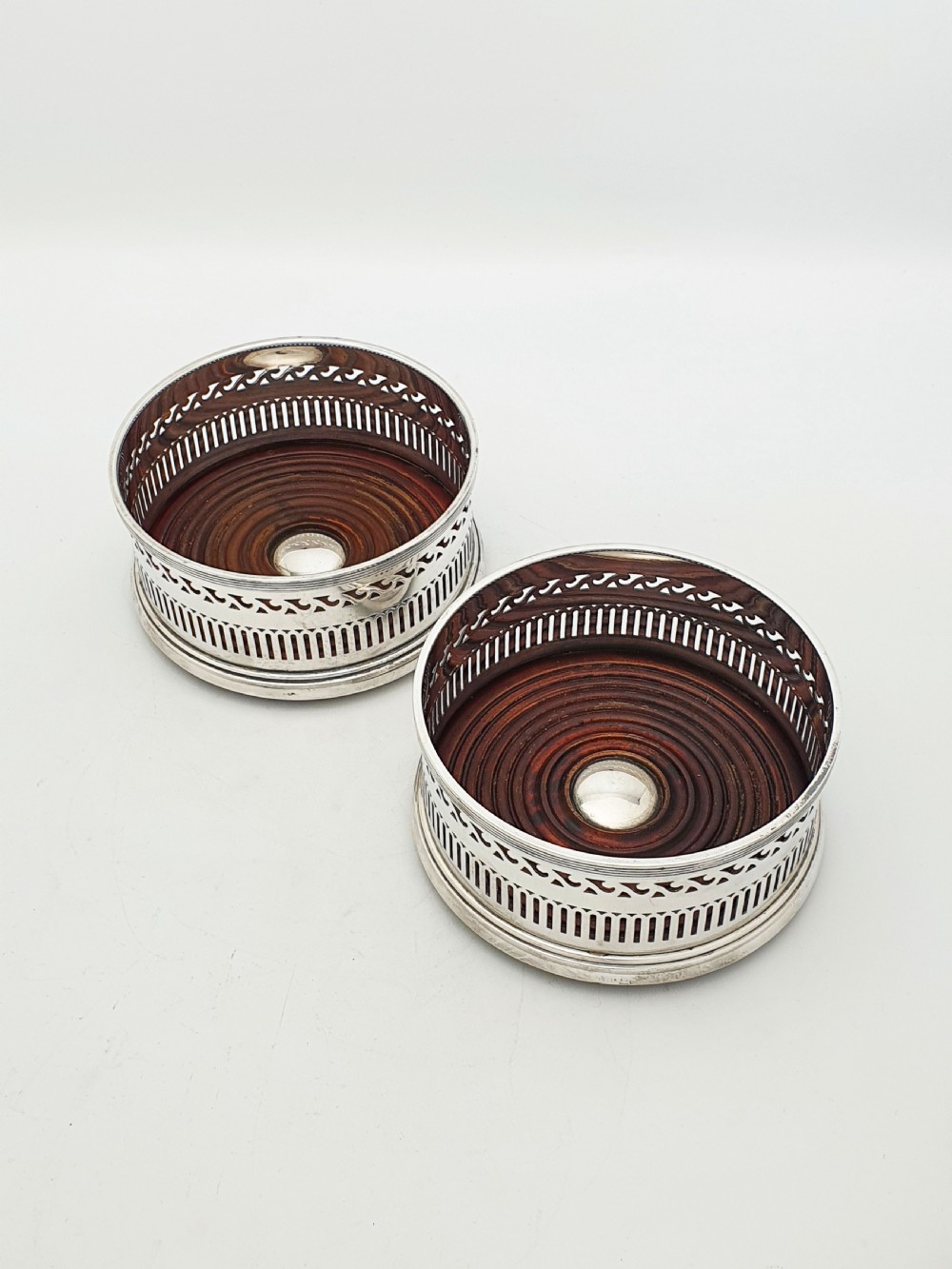 A pair of circular silver plated wine coasters featuring pierced fretwork sides, turned mahogany bases with central silver bosses, and dark felt underneath.