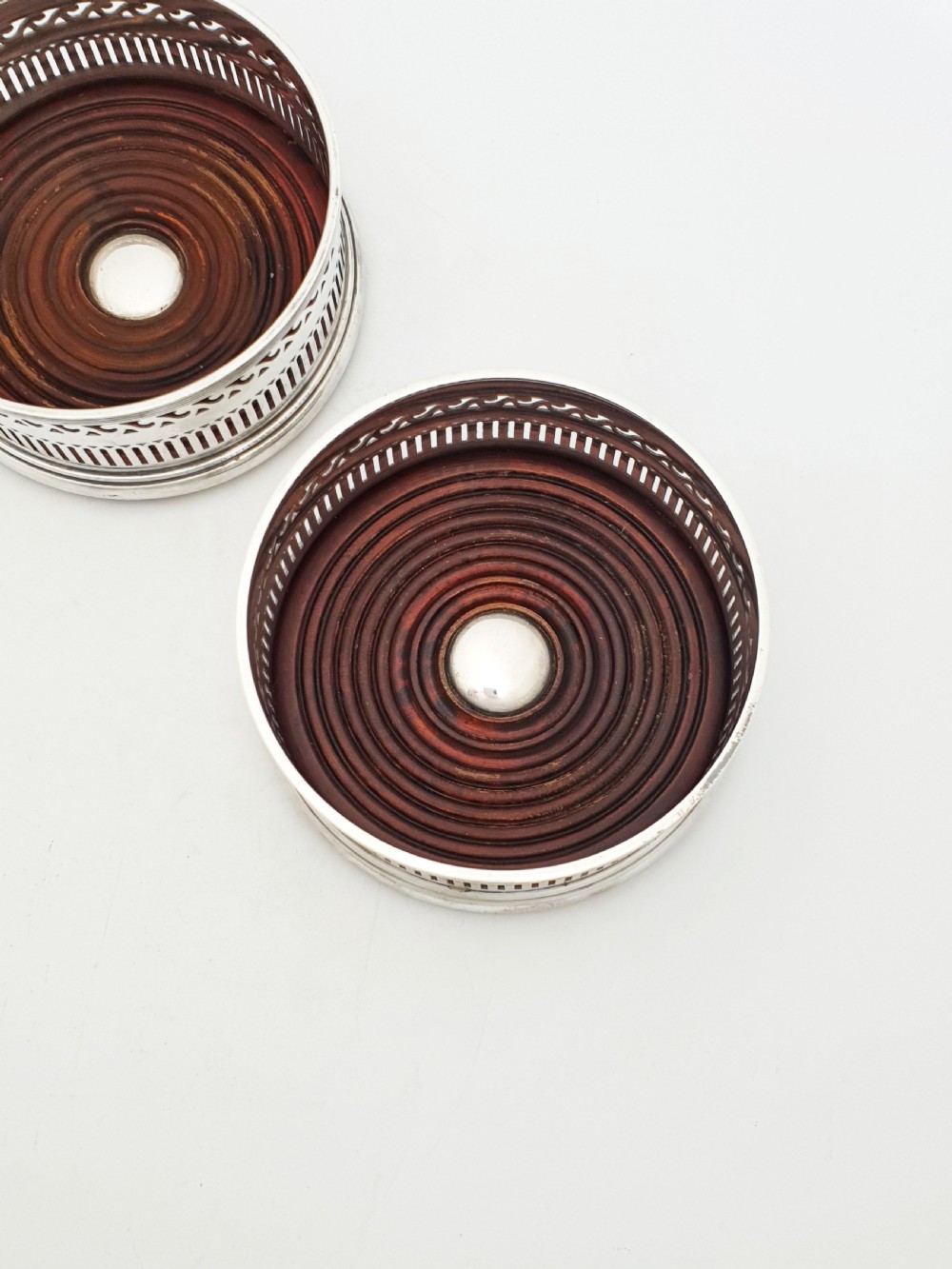 A pair of circular silver plated wine coasters featuring pierced fretwork sides, turned mahogany bases with central silver bosses, and dark felt underneath. View II