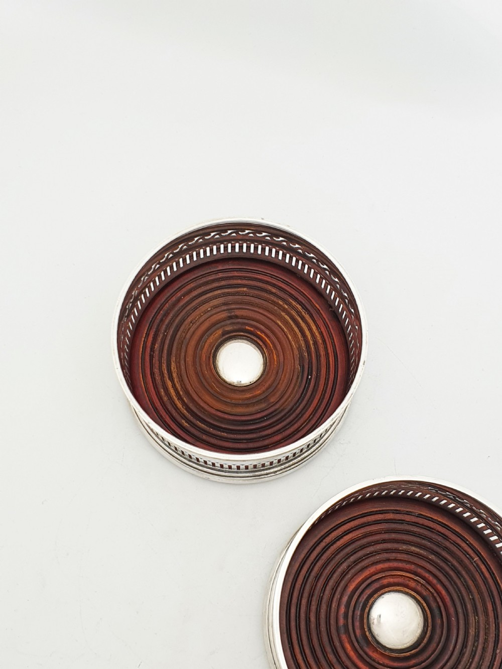 A pair of circular silver plated wine coasters featuring pierced fretwork sides, turned mahogany bases with central silver bosses, and dark felt underneath. View III