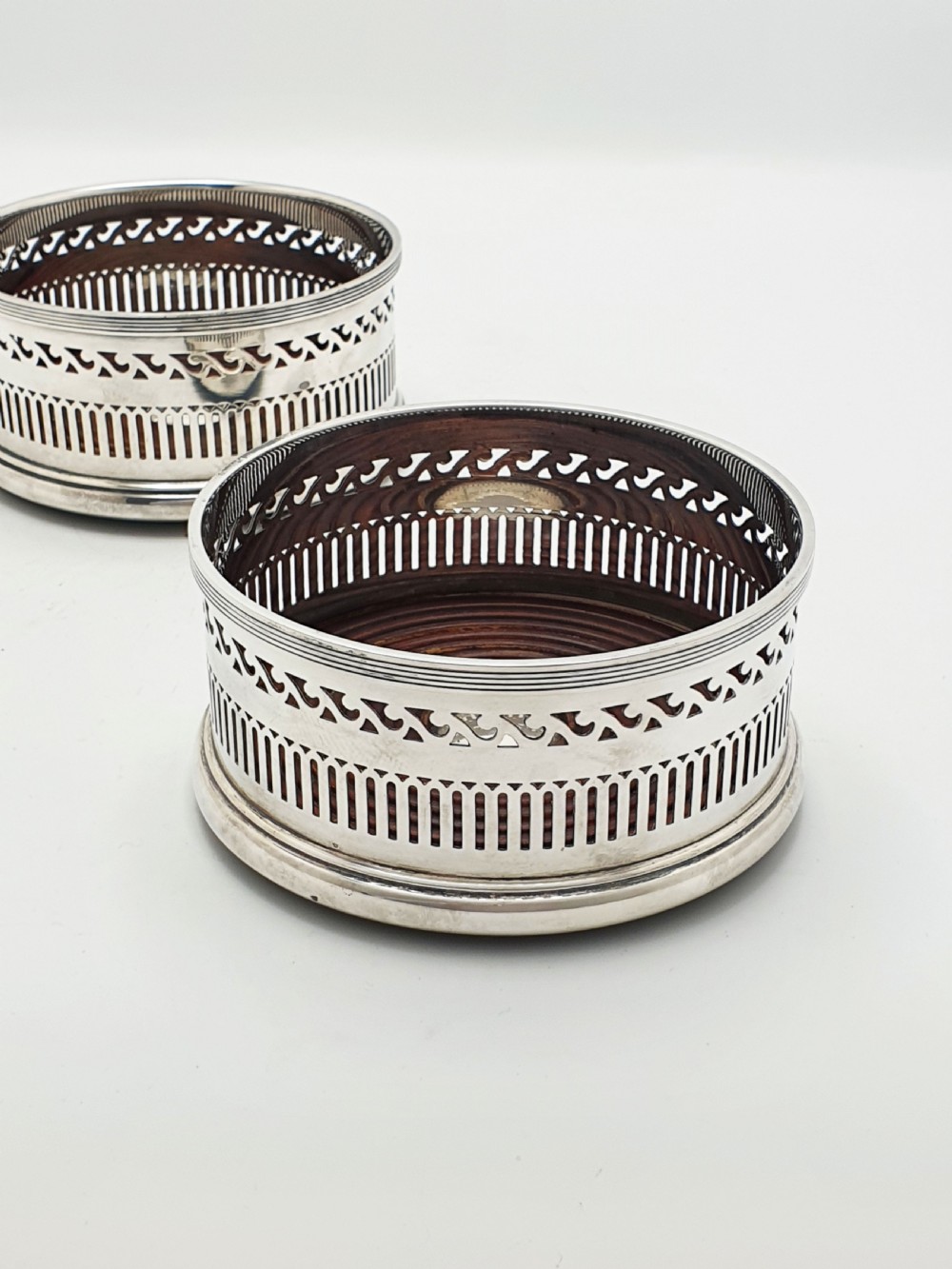 A pair of circular silver plated wine coasters featuring pierced fretwork sides, turned mahogany bases with central silver bosses, and dark felt underneath. View IV