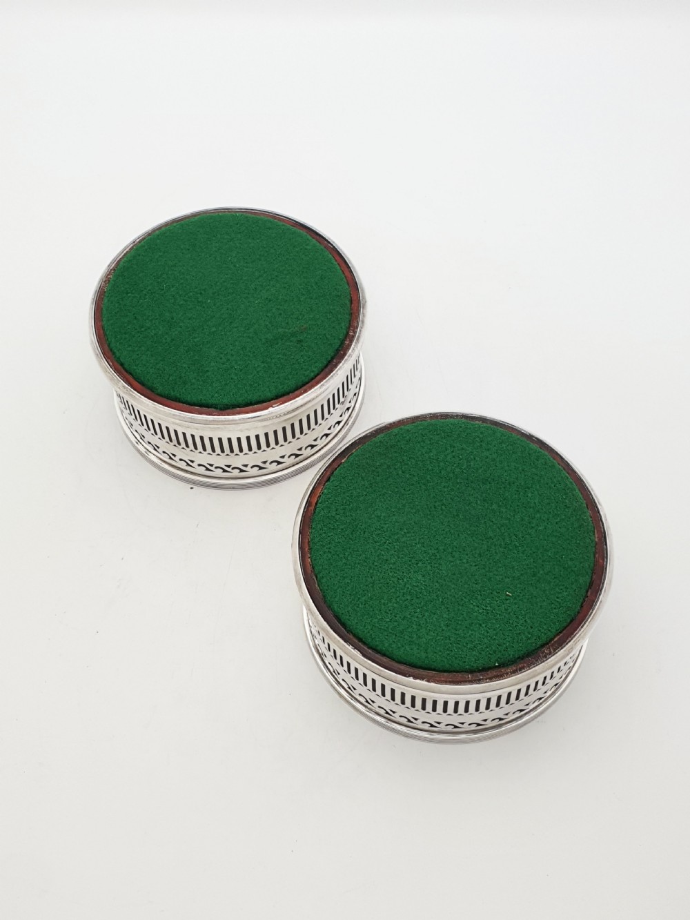 A pair of circular silver plated wine coasters featuring pierced fretwork sides, turned mahogany bases with central silver bosses, and dark felt underneath. View VI