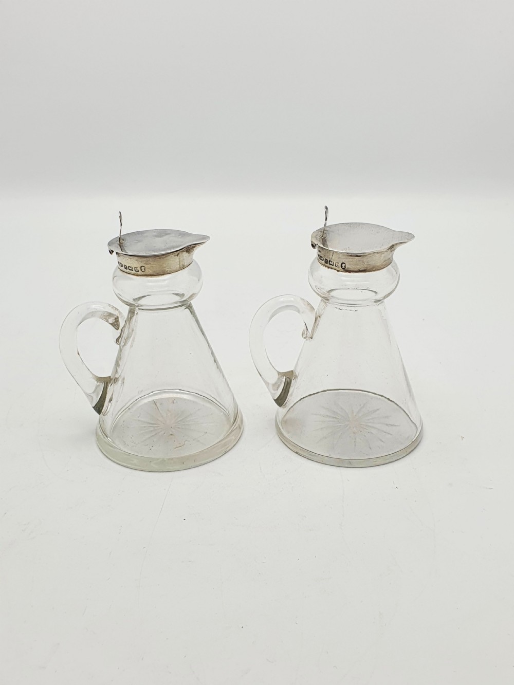 A pair of clear glass conical whiskey noggins with sterling silver collars, hinged lids, and applied glass loop handles.