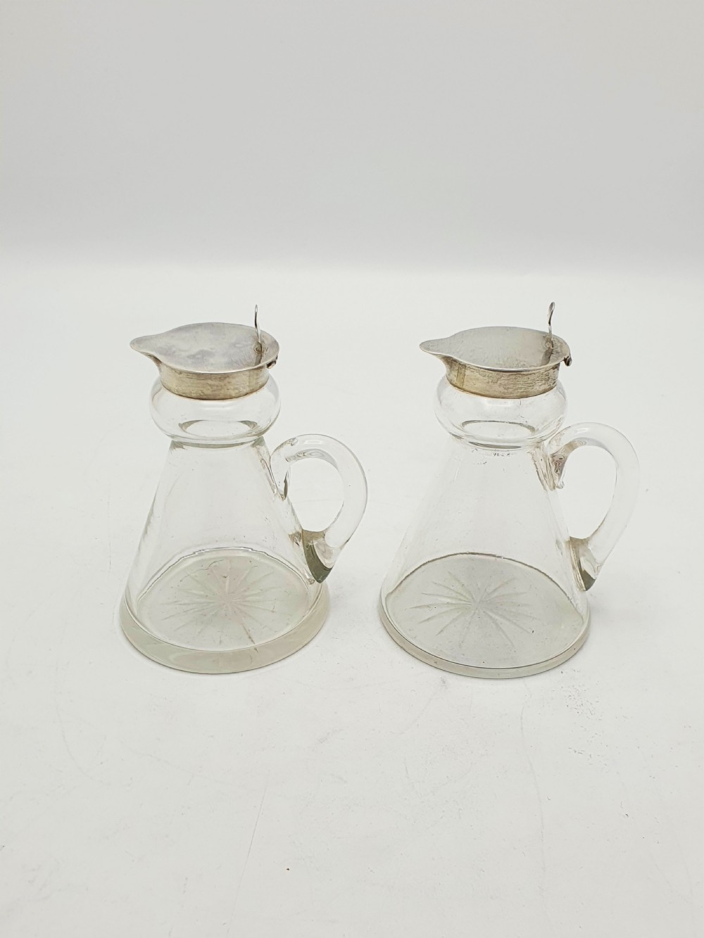 A pair of clear glass conical whiskey noggins with sterling silver collars, hinged lids, and applied glass loop handles. View IX