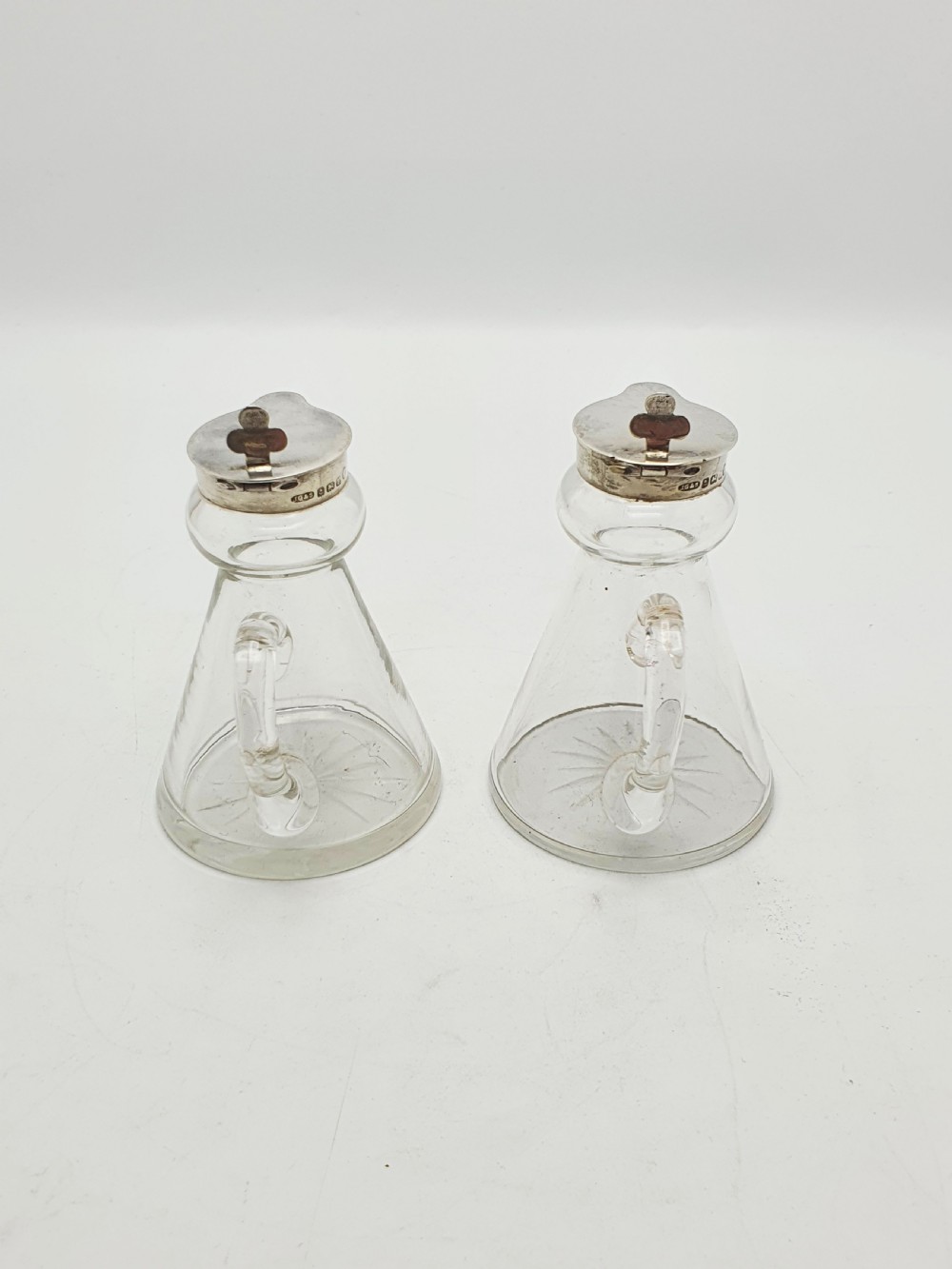 A pair of clear glass conical whiskey noggins with sterling silver collars, hinged lids, and applied glass loop handles. View X