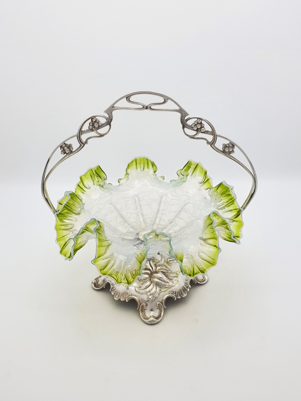 An Art Nouveau silver-plated centrepiece frame with an arched handle holding a frilled green-rimmed vaseline glass dish with a crackle texture.