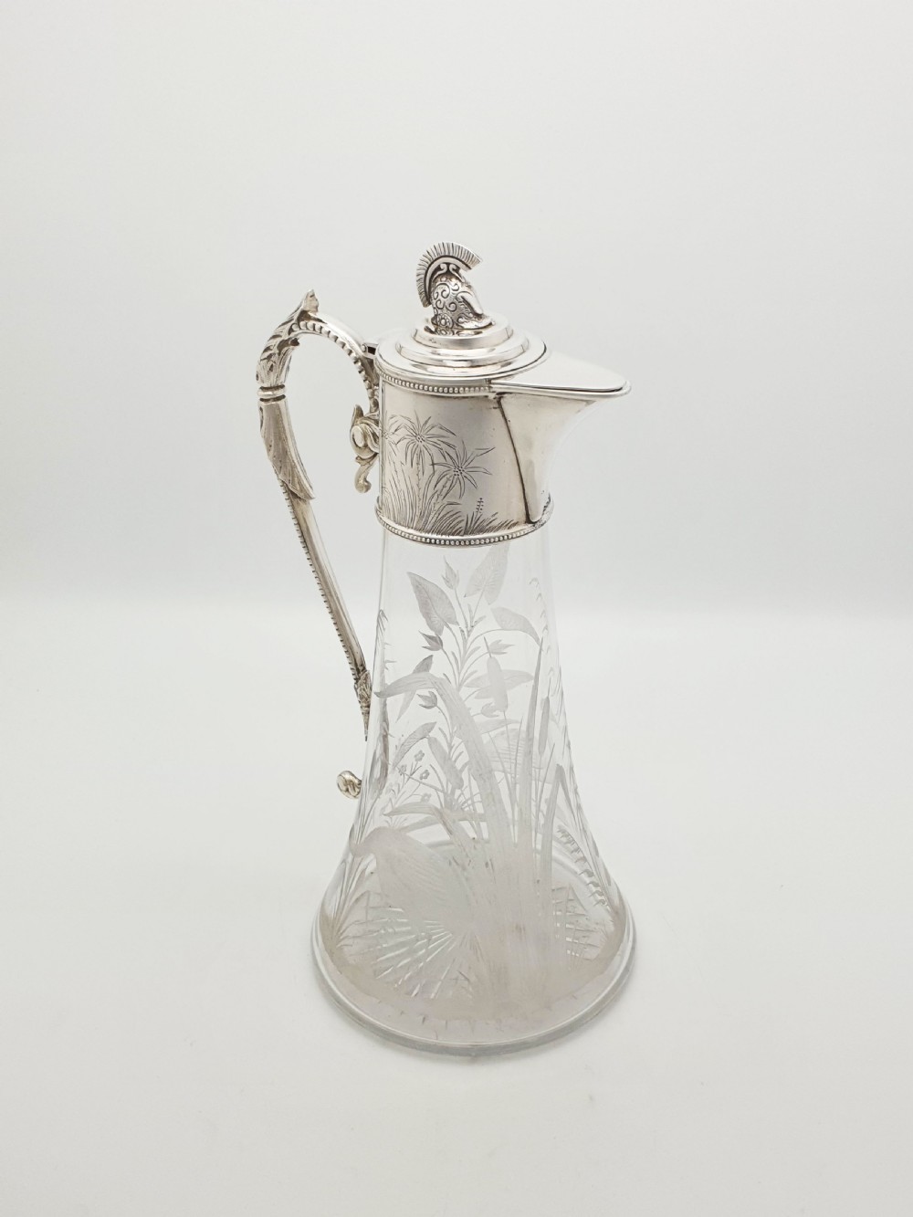 A Victorian etched clear glass claret jug featuring botanical engravings and a silver-plated handle, spout, and helmet finial.