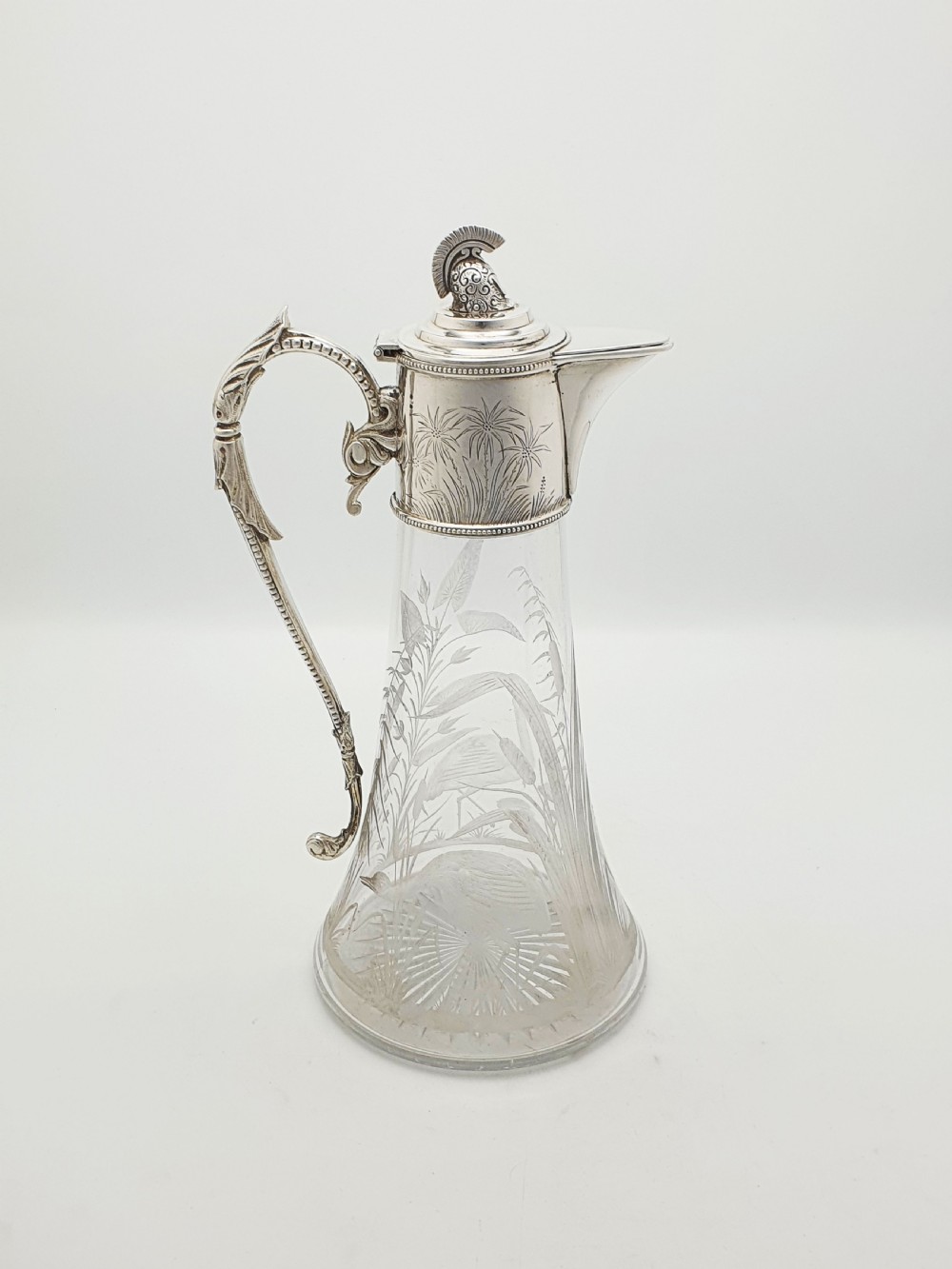 A Victorian etched clear glass claret jug featuring botanical engravings and a silver-plated handle, spout, and helmet finial. View VI
