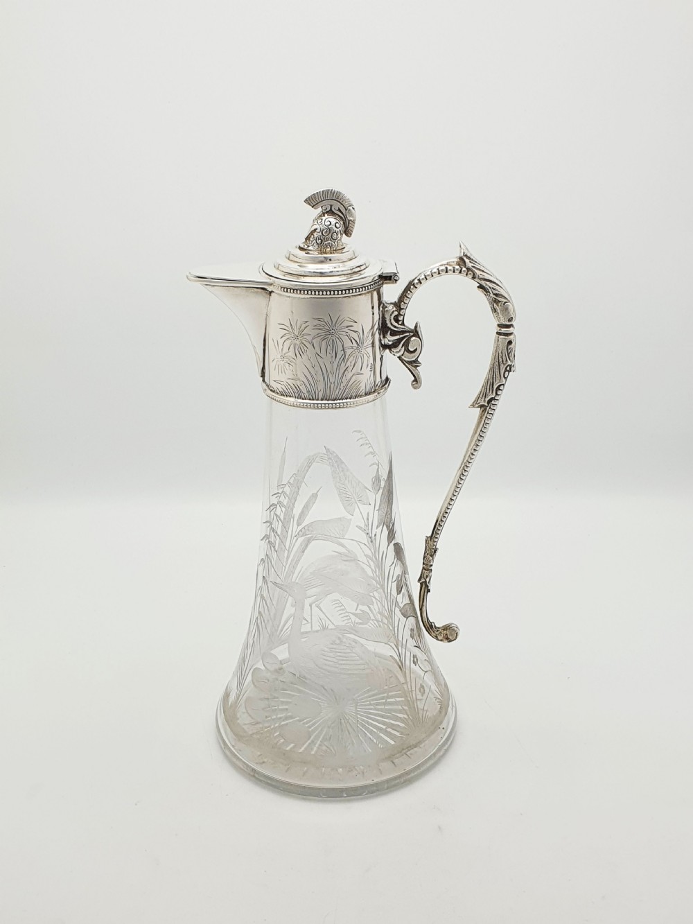 A Victorian etched clear glass claret jug featuring botanical engravings and a silver-plated handle, spout, and helmet finial. View VIII