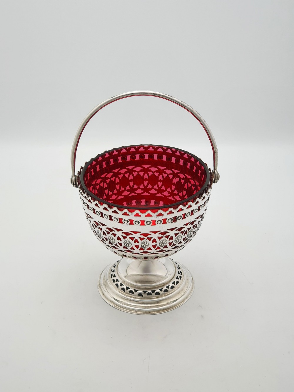 An Edwardian silver-plated footed basket featuring pierced geometric fretwork, a swing handle, and a removable ruby red glass liner.