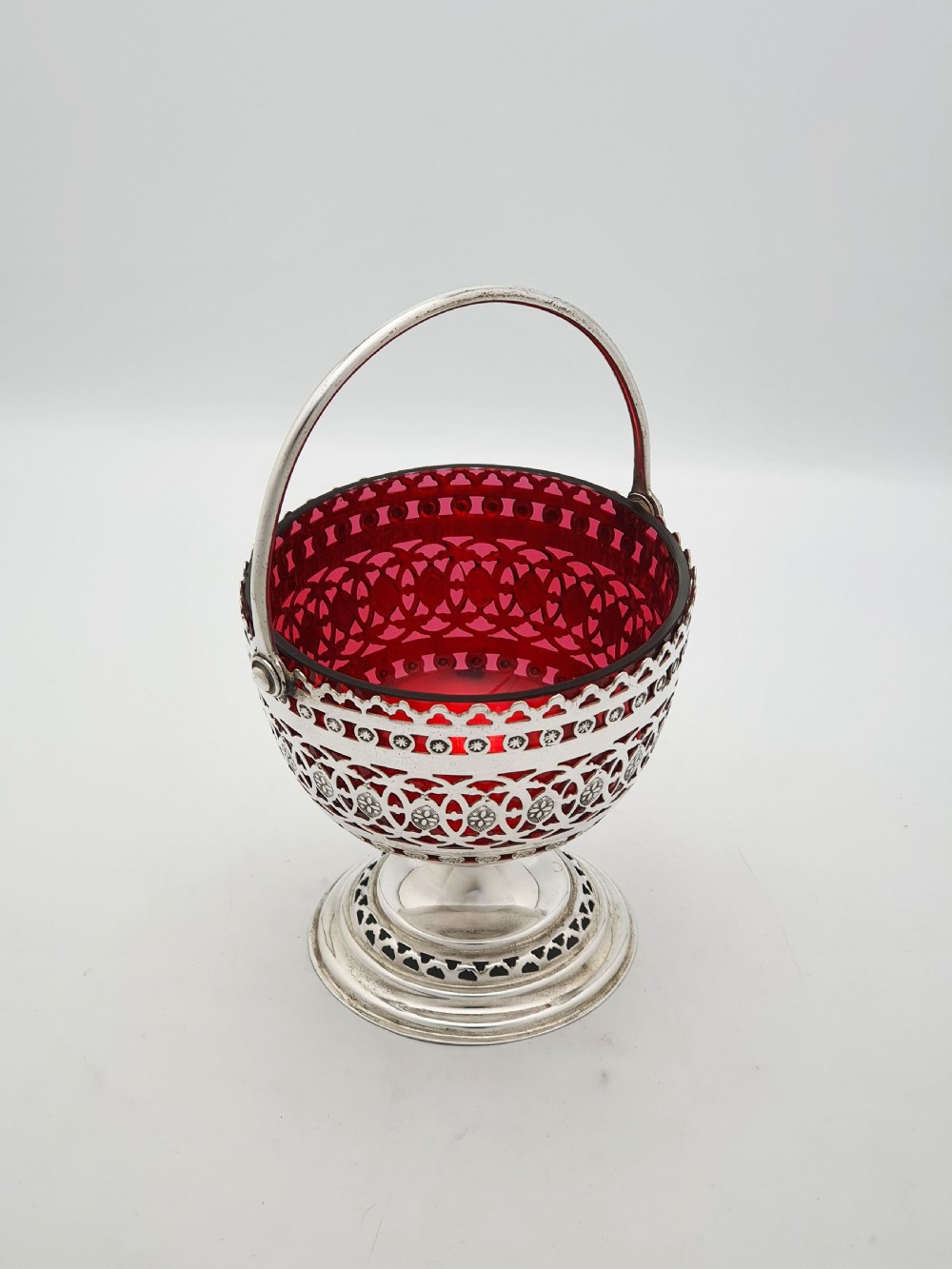 An Edwardian silver-plated footed basket featuring pierced geometric fretwork, a swing handle, and a removable ruby red glass liner. View II