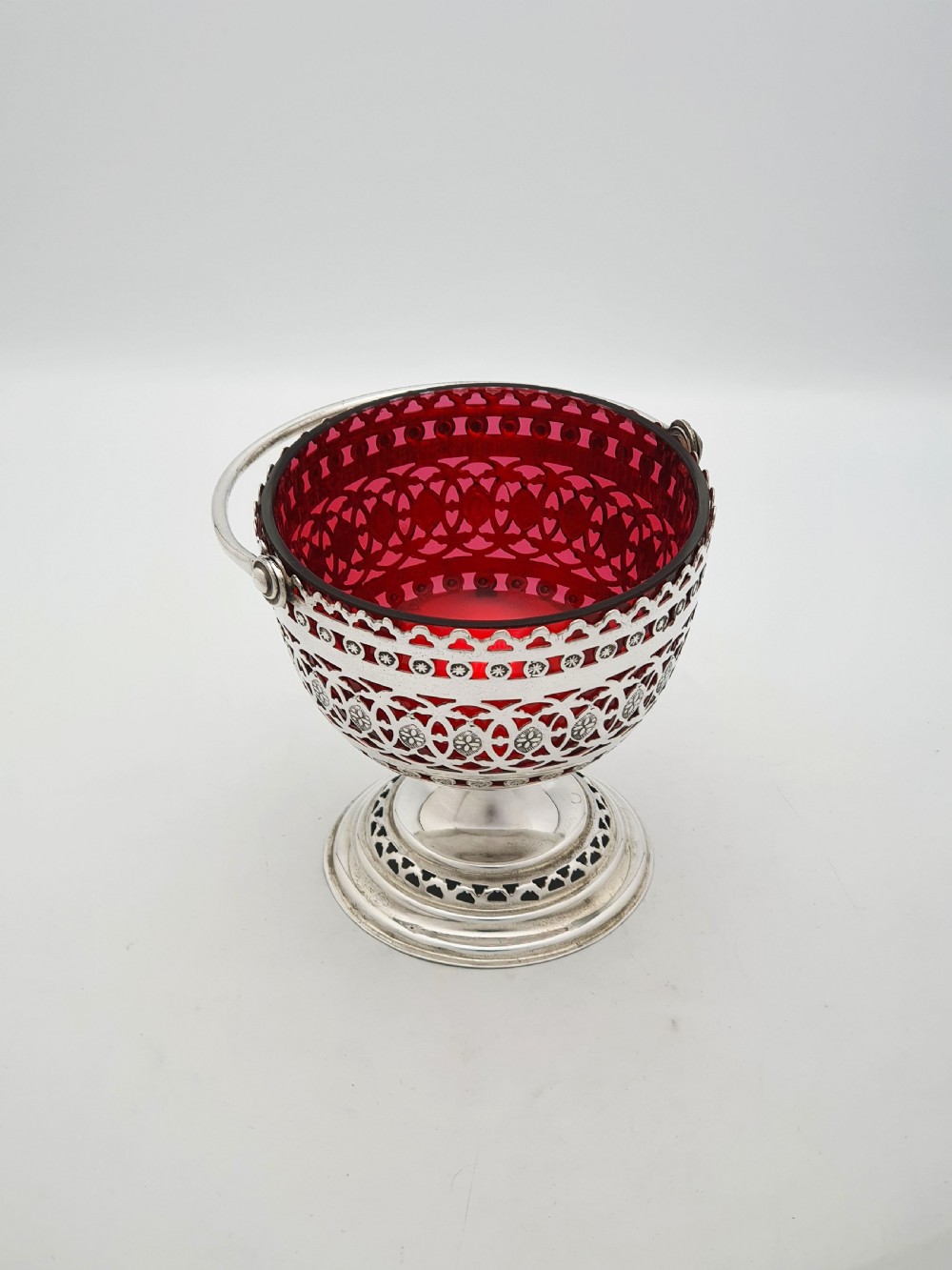 An Edwardian silver-plated footed basket featuring pierced geometric fretwork, a swing handle, and a removable ruby red glass liner. View III