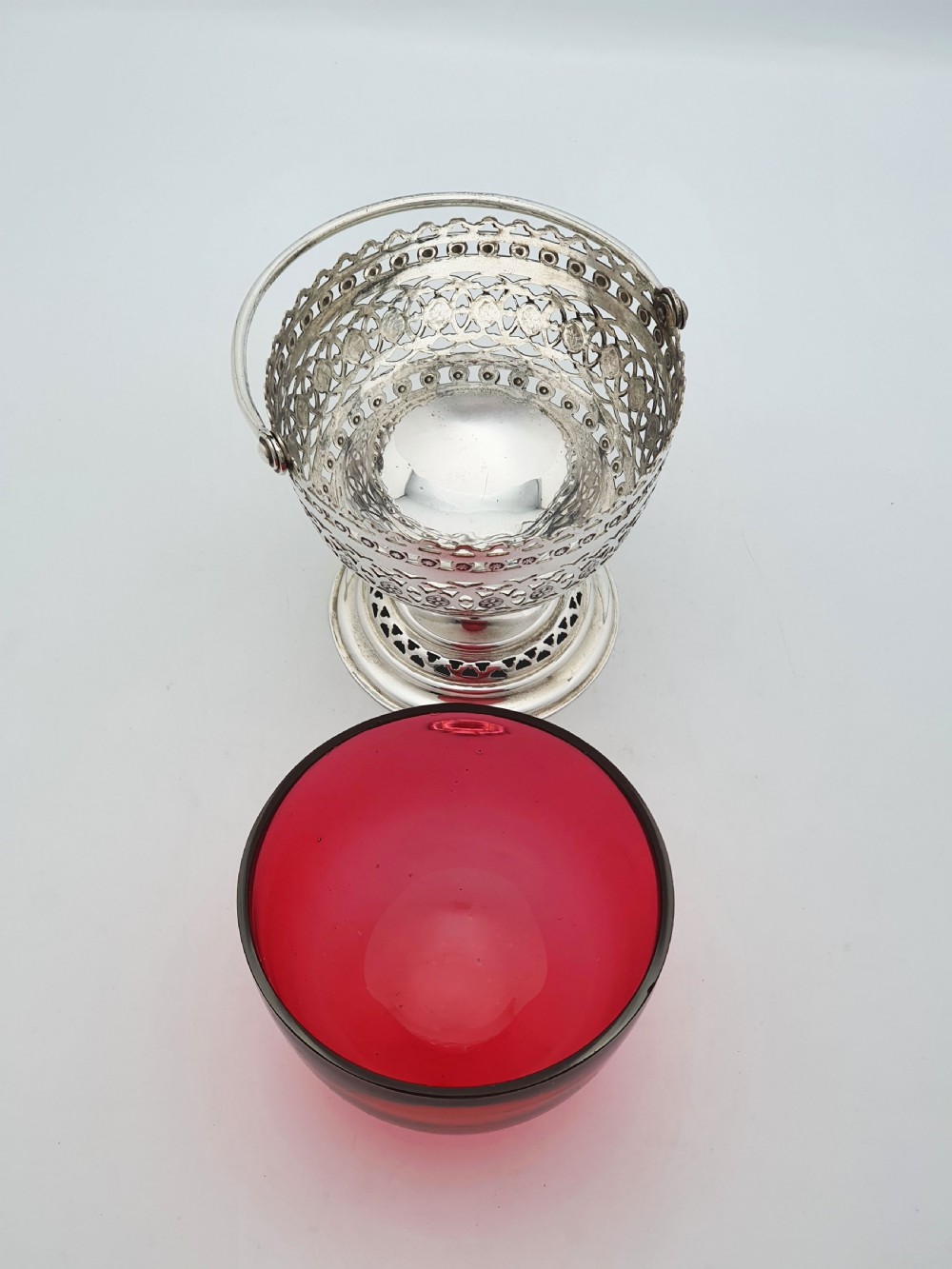 An Edwardian silver-plated footed basket featuring pierced geometric fretwork, a swing handle, and a removable ruby red glass liner. View V