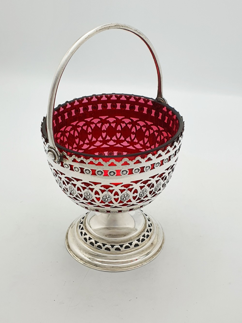 An Edwardian silver-plated footed basket featuring pierced geometric fretwork, a swing handle, and a removable ruby red glass liner. View VI