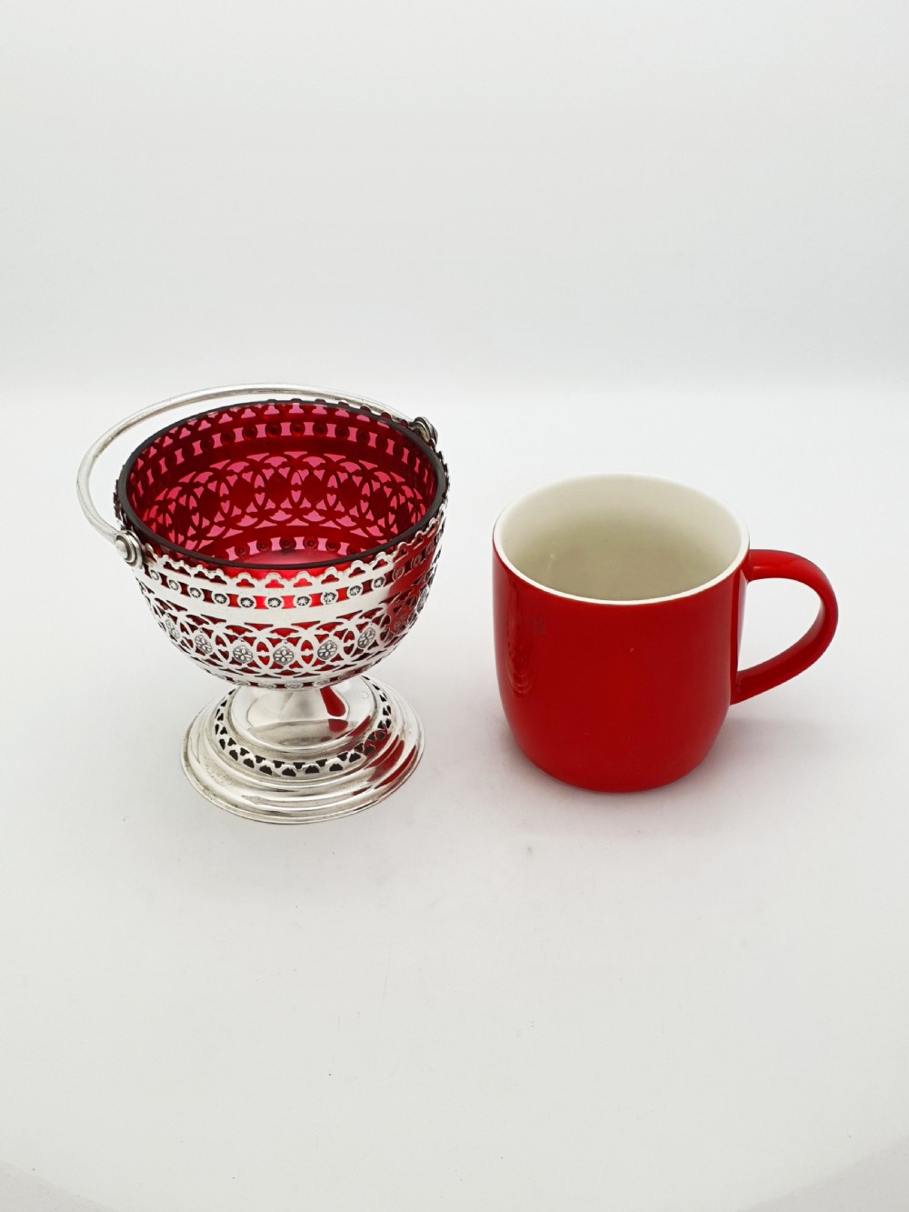 An Edwardian silver-plated footed basket featuring pierced geometric fretwork, a swing handle, and a removable ruby red glass liner. View VII