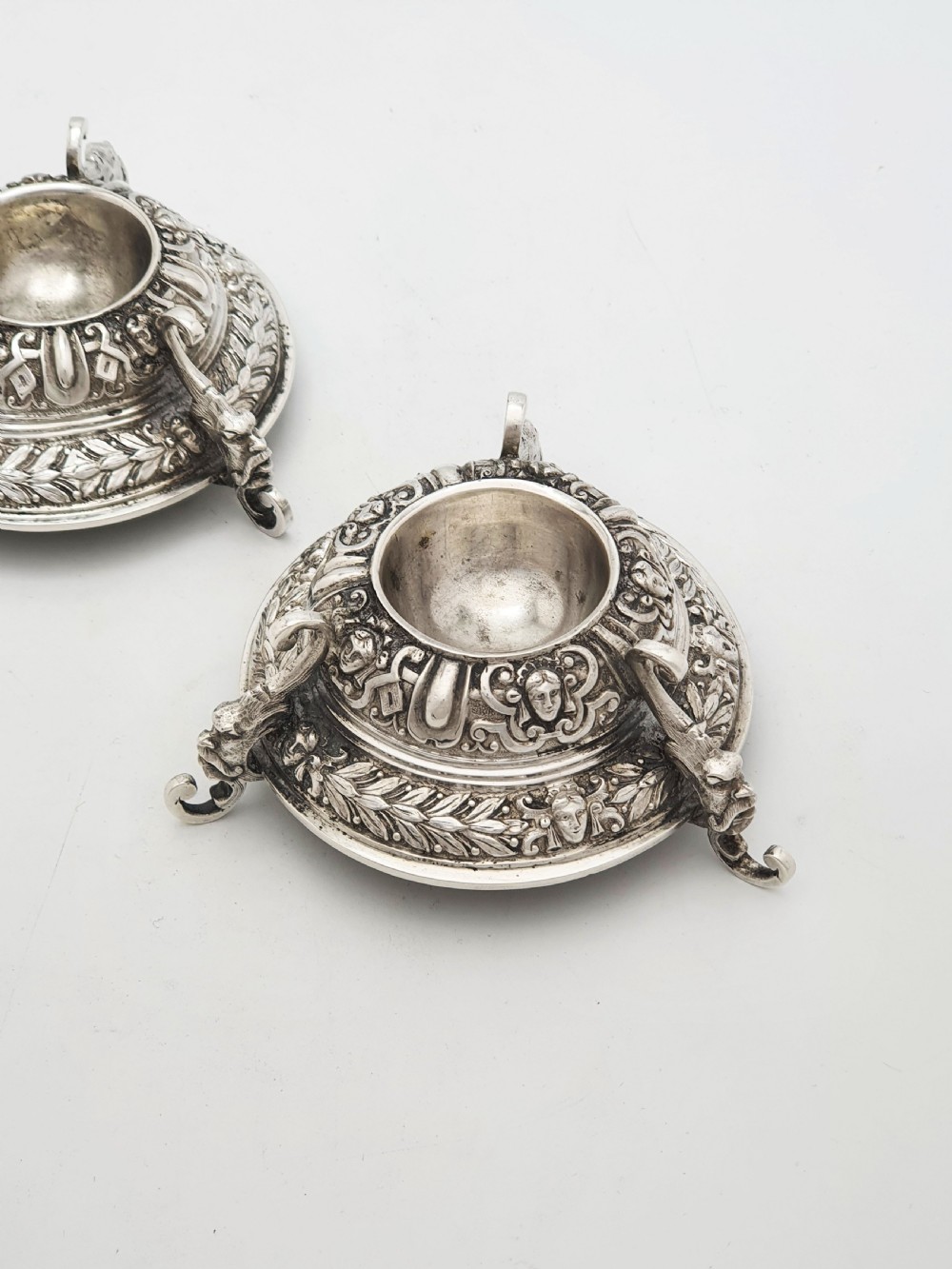 A pair of Victorian sterling silver salt cellars decorated with chased classical masks, foliate wreaths, and tripod feet.