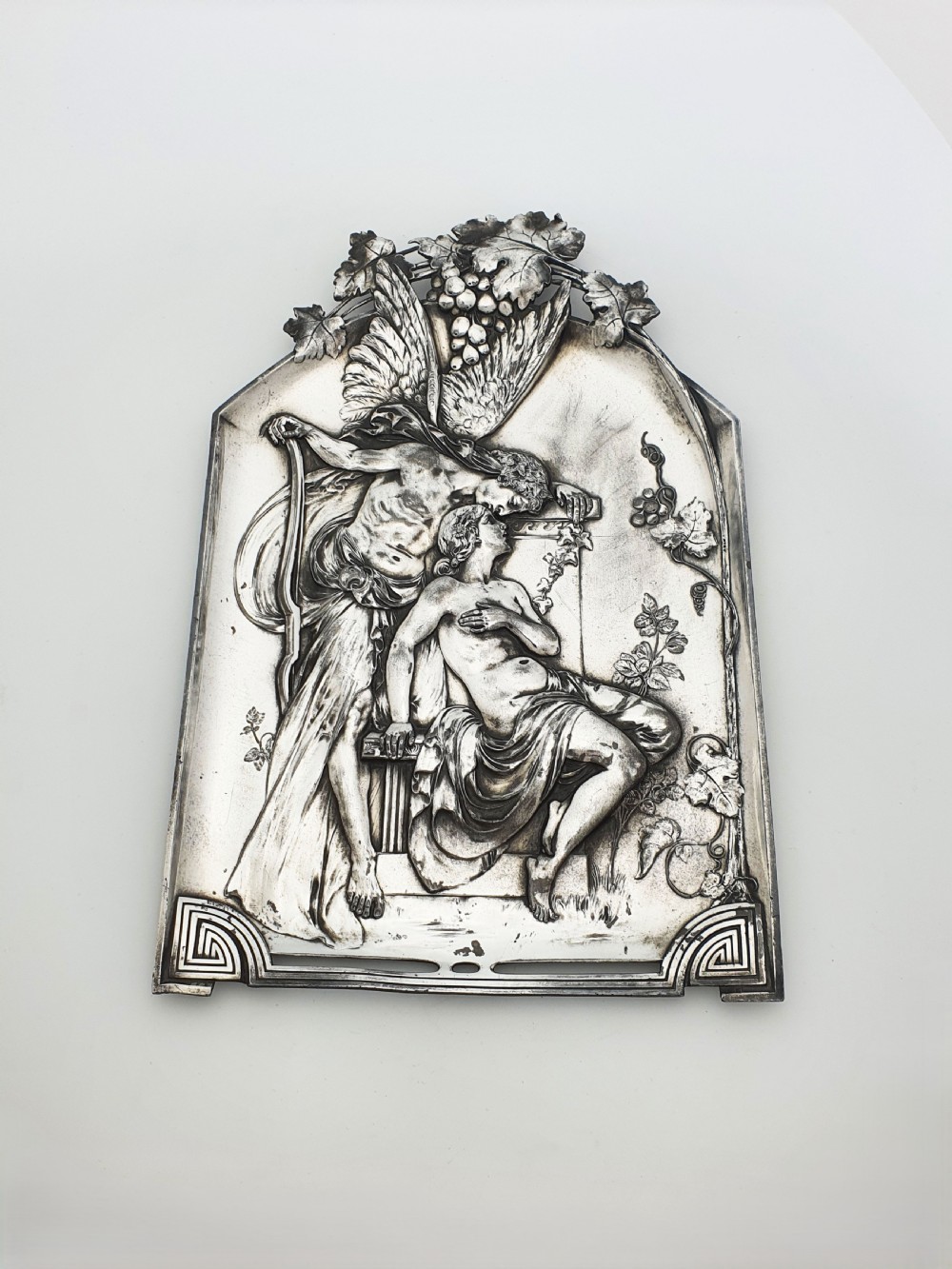 An arched silver-plated metal relief wall plaque featuring a classical mythological figure scene framed by grapevines and geometric motifs.