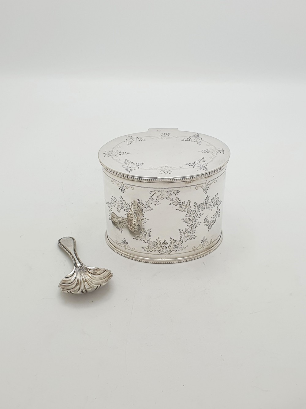 An oval silver-plated tea caddy featuring engraved foliate garlands on the body and lid, accompanied by a small shell-shaped spoon.