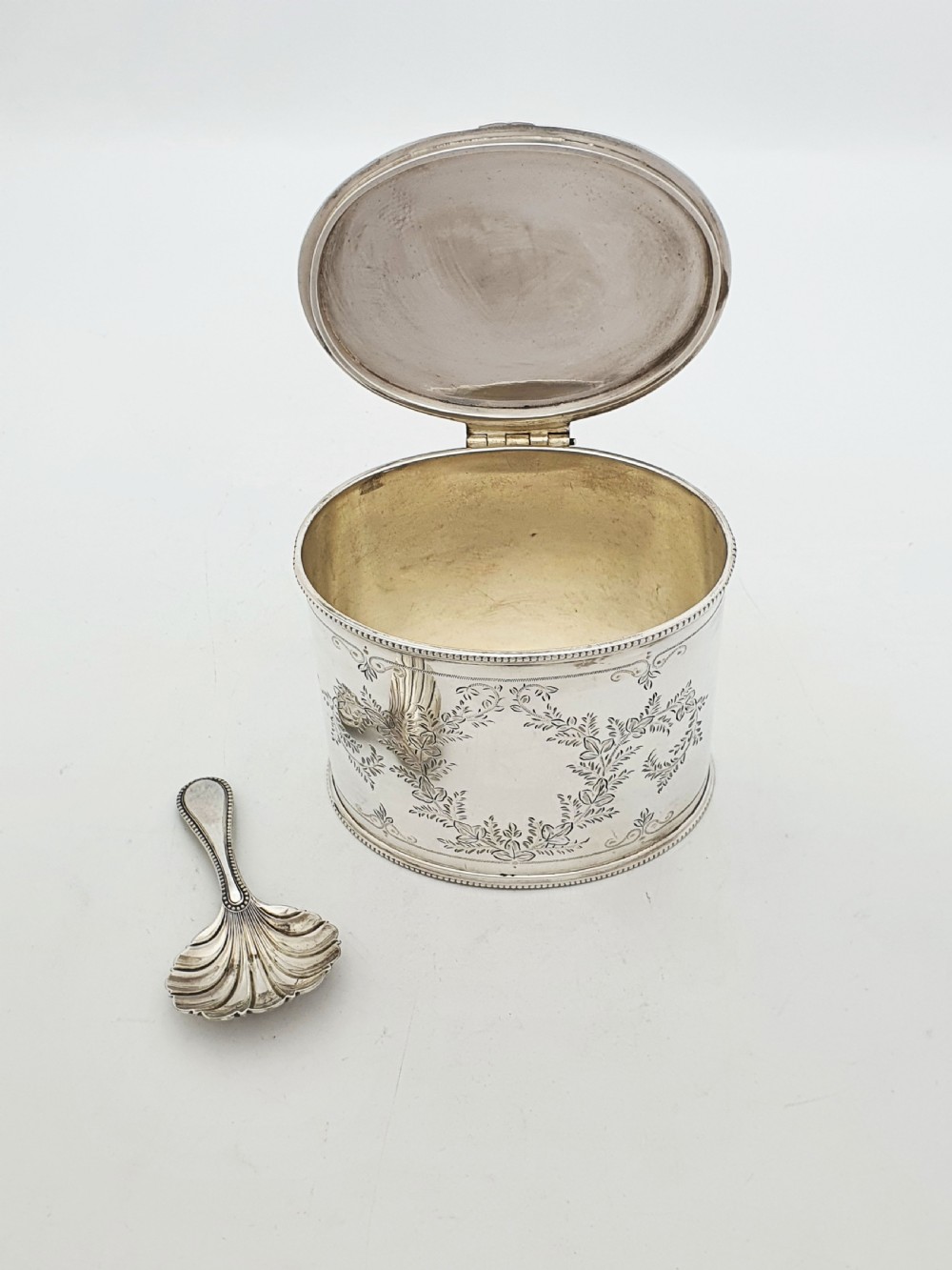 An oval silver-plated tea caddy featuring engraved foliate garlands on the body and lid, accompanied by a small shell-shaped spoon. View II
