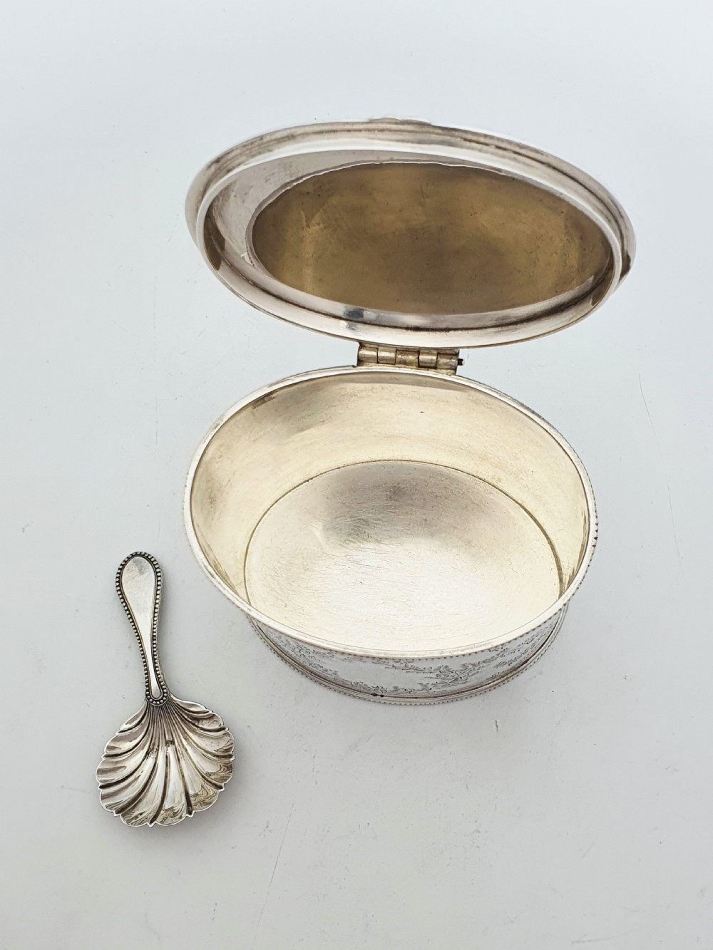 An oval silver-plated tea caddy featuring engraved foliate garlands on the body and lid, accompanied by a small shell-shaped spoon. View III