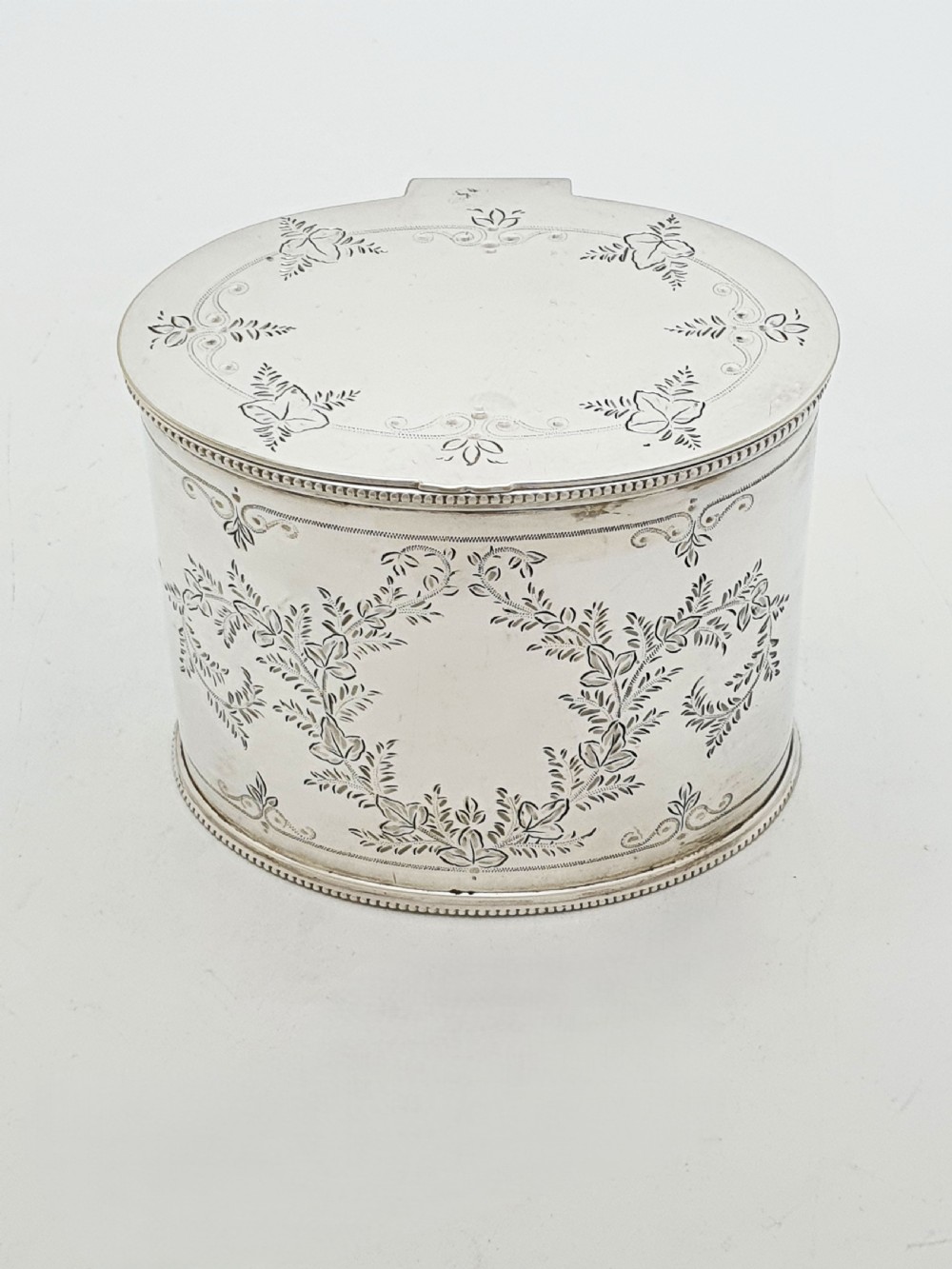 An oval silver-plated tea caddy featuring engraved foliate garlands on the body and lid, accompanied by a small shell-shaped spoon. View IV