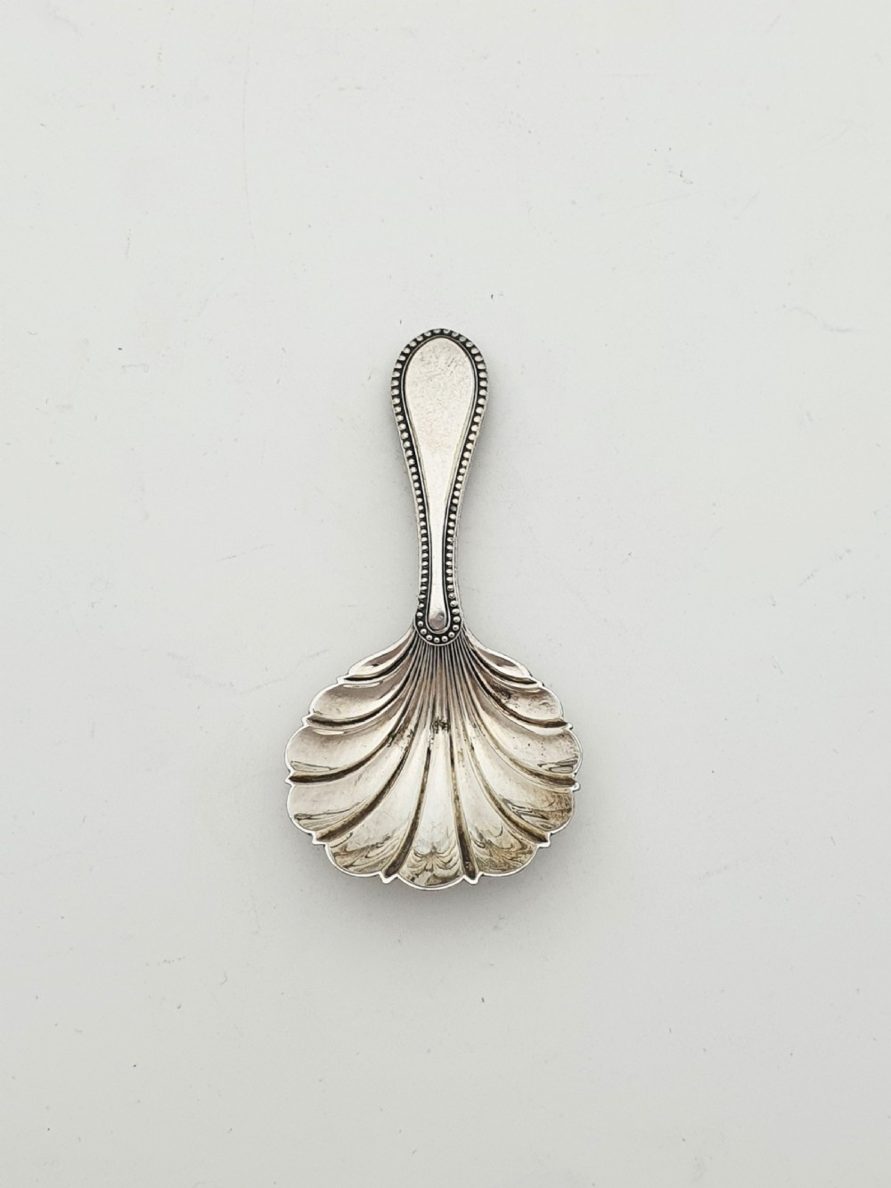 An oval silver-plated tea caddy featuring engraved foliate garlands on the body and lid, accompanied by a small shell-shaped spoon. View IX