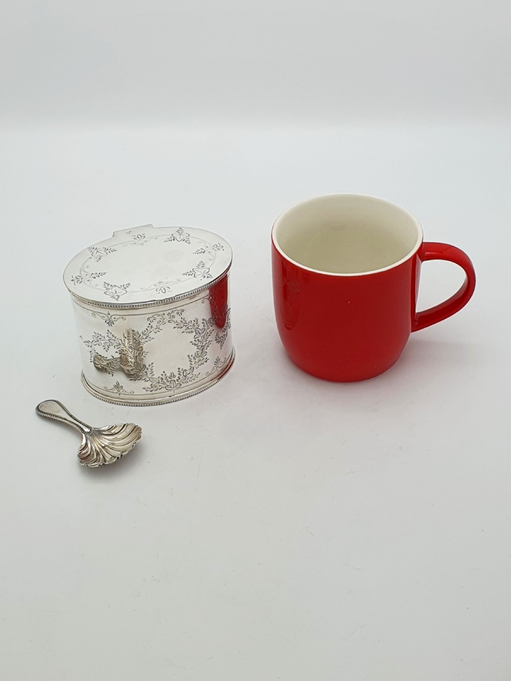 An oval silver-plated tea caddy featuring engraved foliate garlands on the body and lid, accompanied by a small shell-shaped spoon. View X