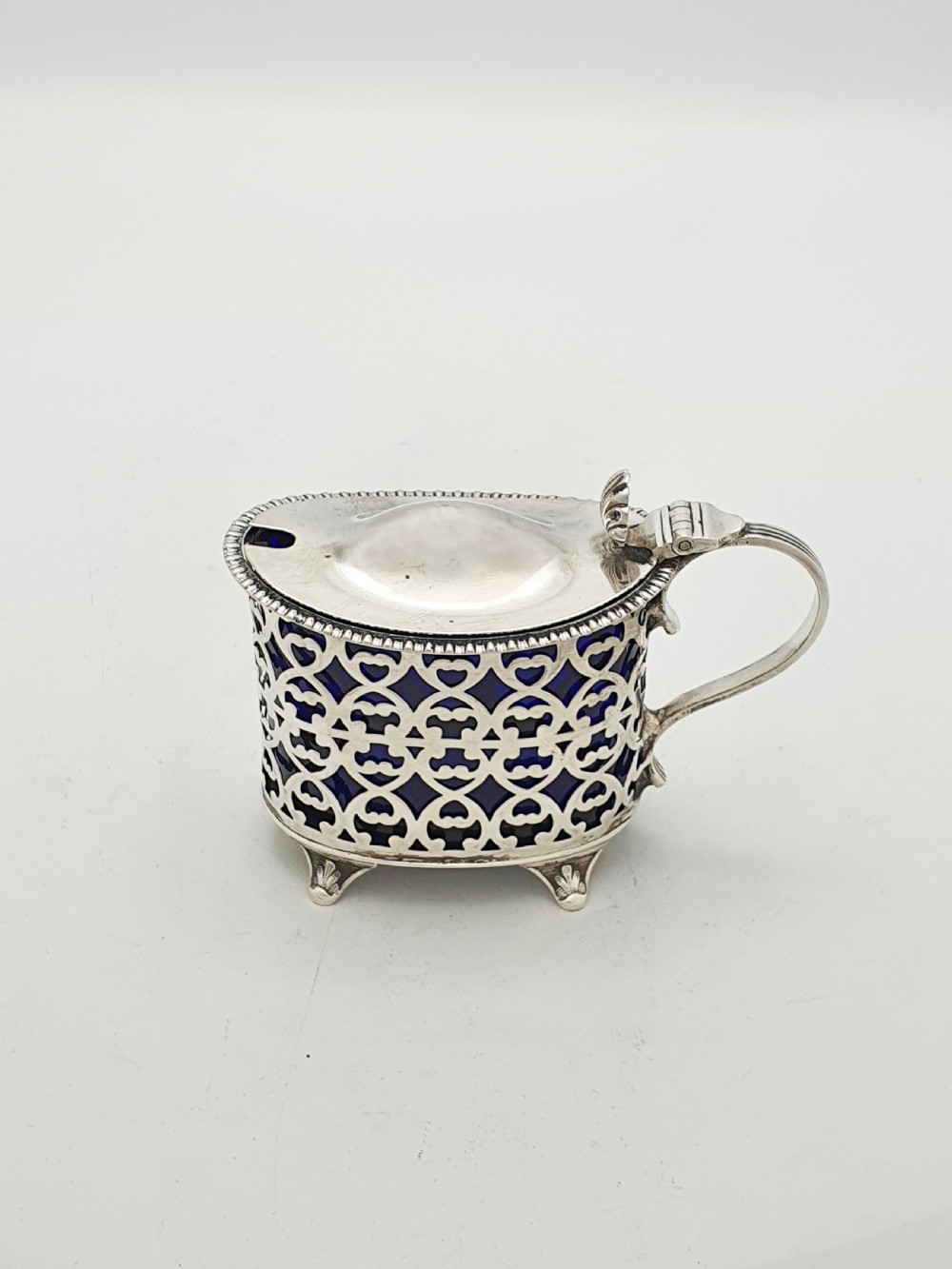 A Victorian sterling silver mustard pot with openwork lattice piercing, a hinged lid, a scroll handle, and a removable cobalt blue glass liner. View IV