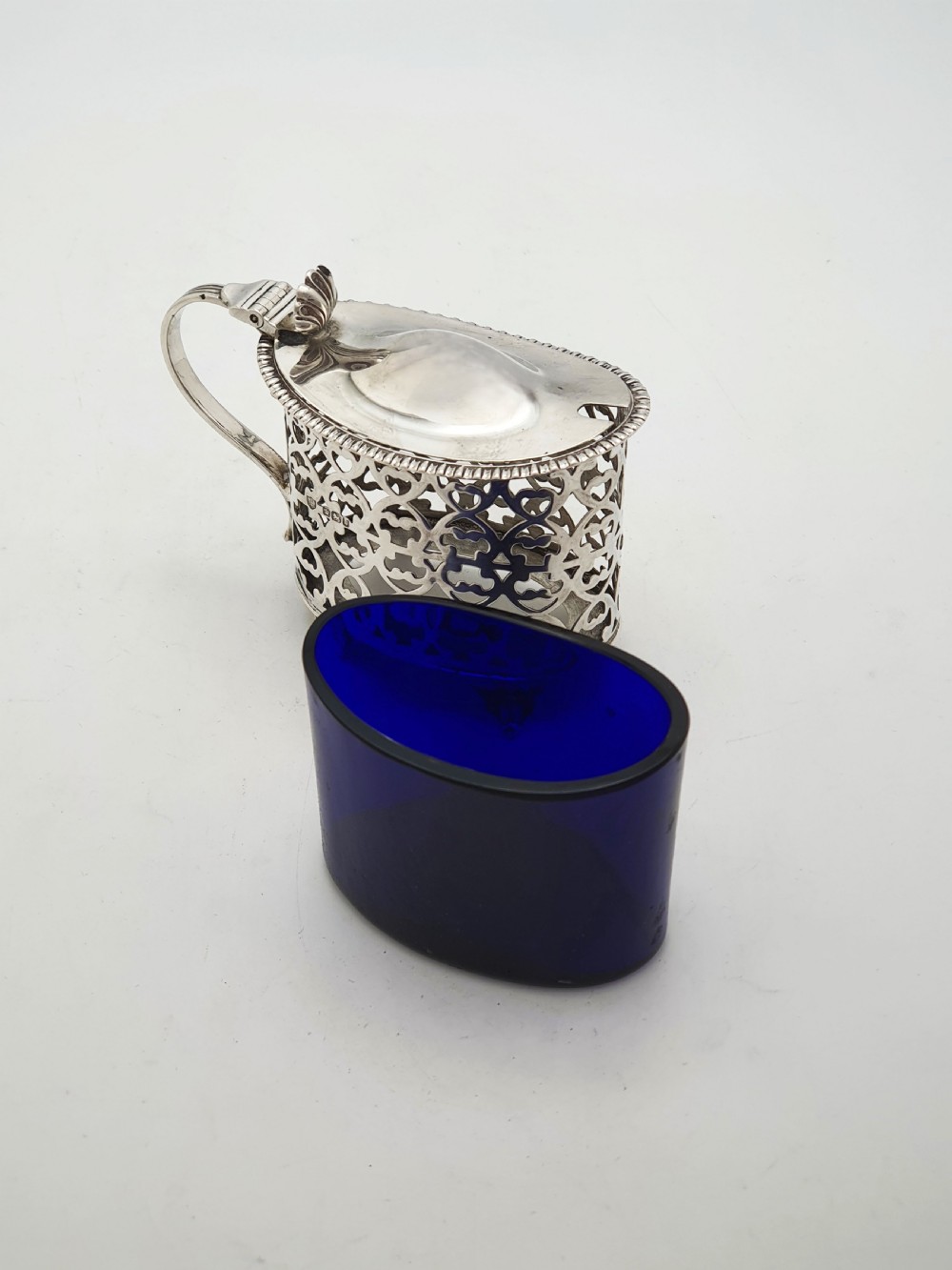 A Victorian sterling silver mustard pot with openwork lattice piercing, a hinged lid, a scroll handle, and a removable cobalt blue glass liner. View VI