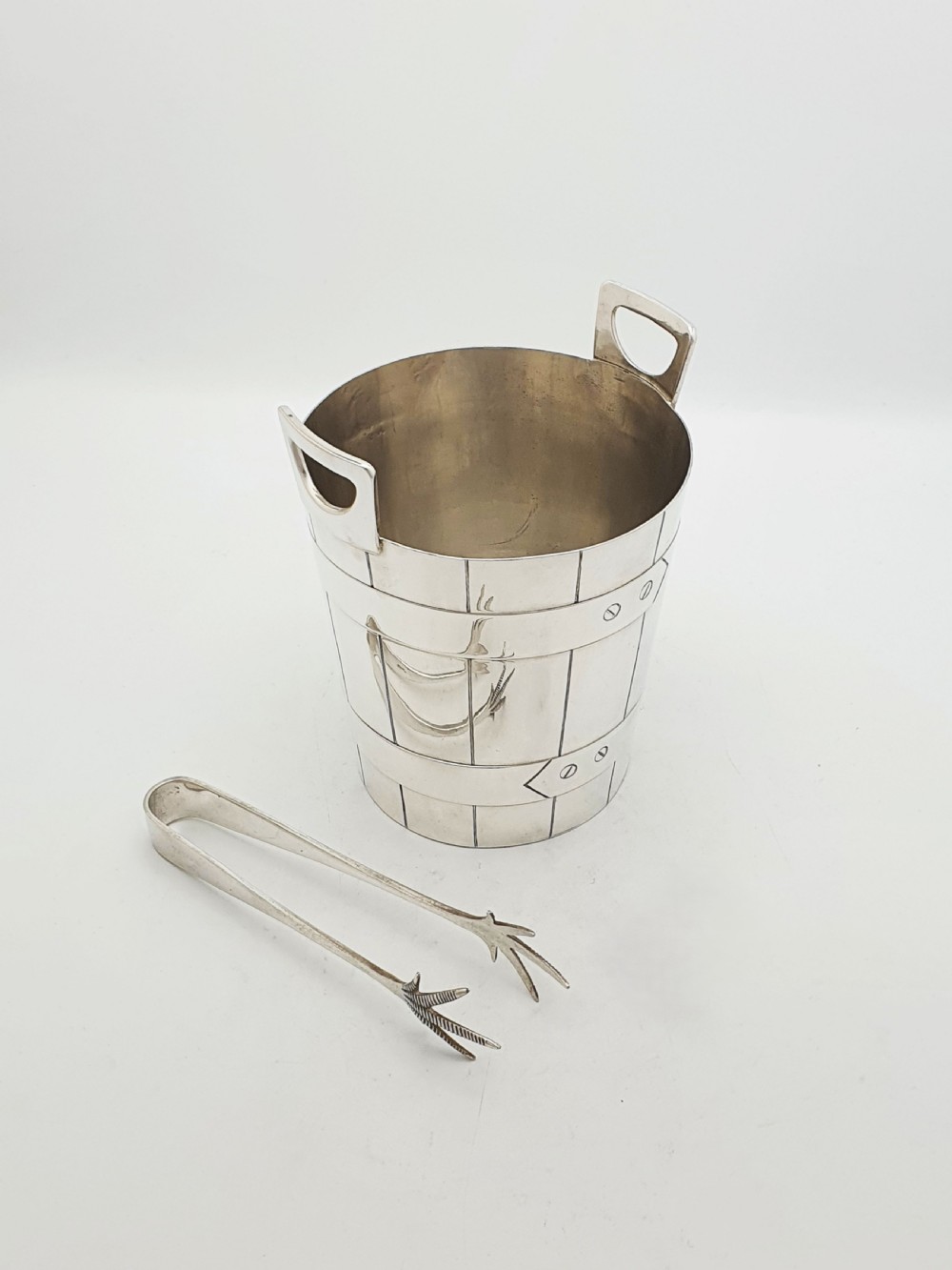 An Edwardian silver-plated ice bucket shaped like a wooden barrel with integrated handles alongside matching claw-foot ice tongs.