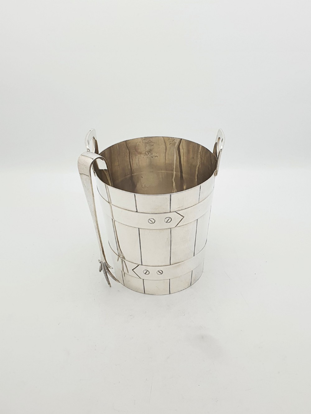 An Edwardian silver-plated ice bucket shaped like a wooden barrel with integrated handles alongside matching claw-foot ice tongs. View II