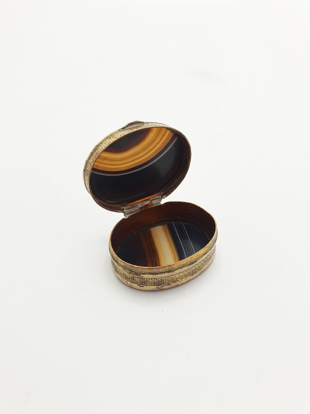 An open small oval Victorian gilt brass box with textured pattern engraving and polished banded agate panels set into the top and bottom.
