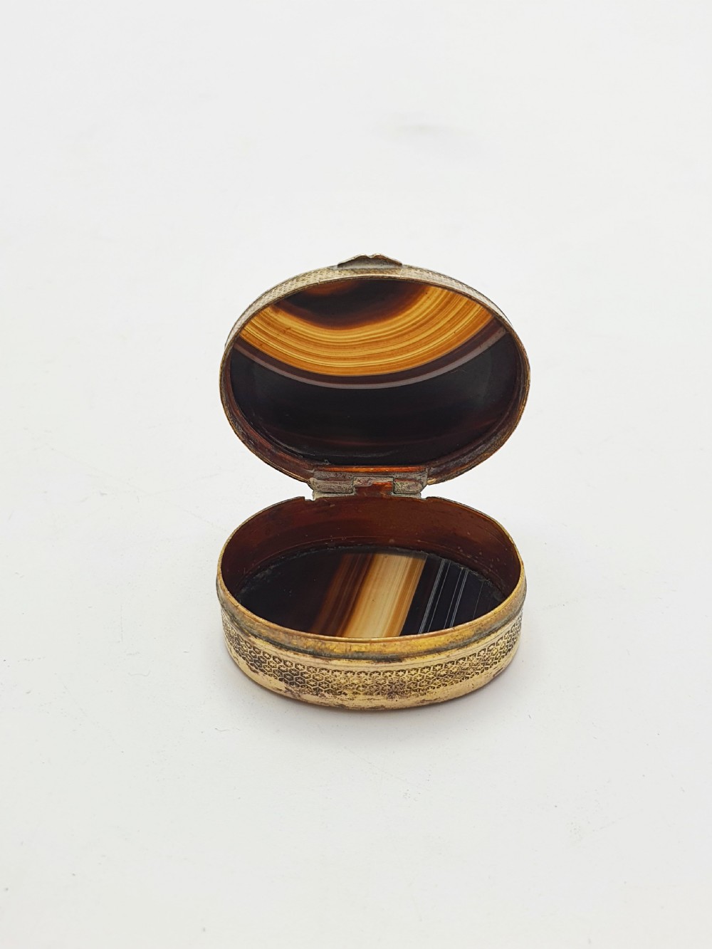 An open small oval Victorian gilt brass box with textured pattern engraving and polished banded agate panels set into the top and bottom. View II