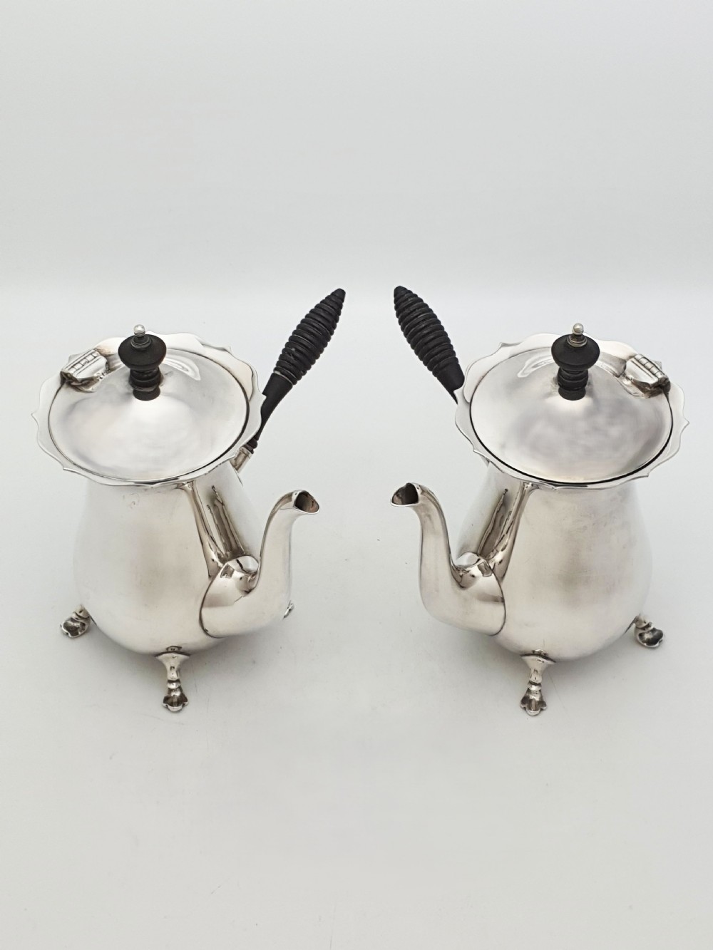 A pair of Edwardian silver cafe au lait pots featuring ribbed wooden side handles, scalloped hinged lids, and carved cabriole feet.