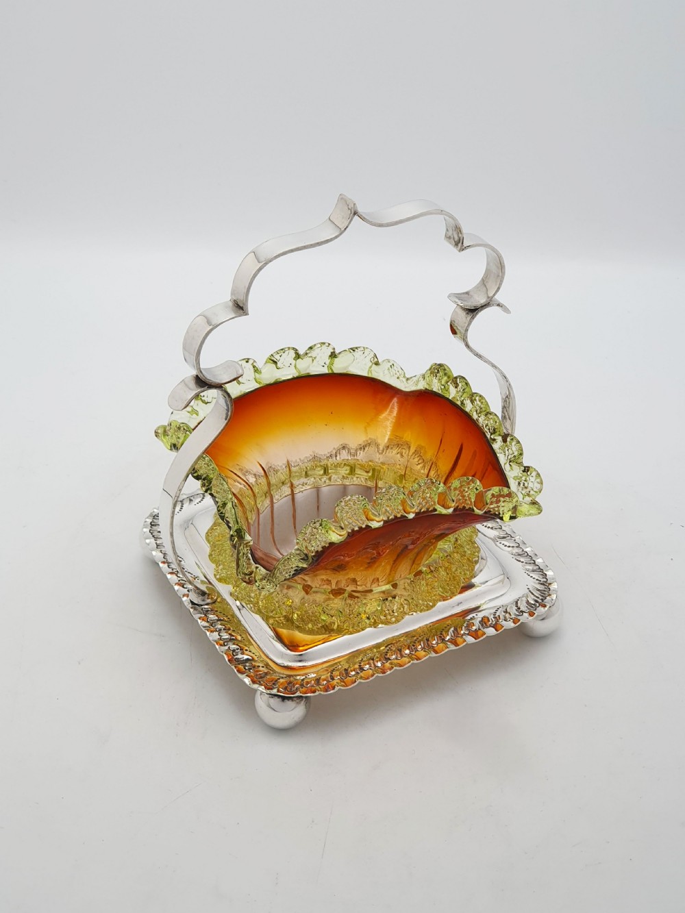 An Edwardian silver-plated stand with a shaped handle and ball feet holding a ruffled amber and yellow glass bonbon dish.