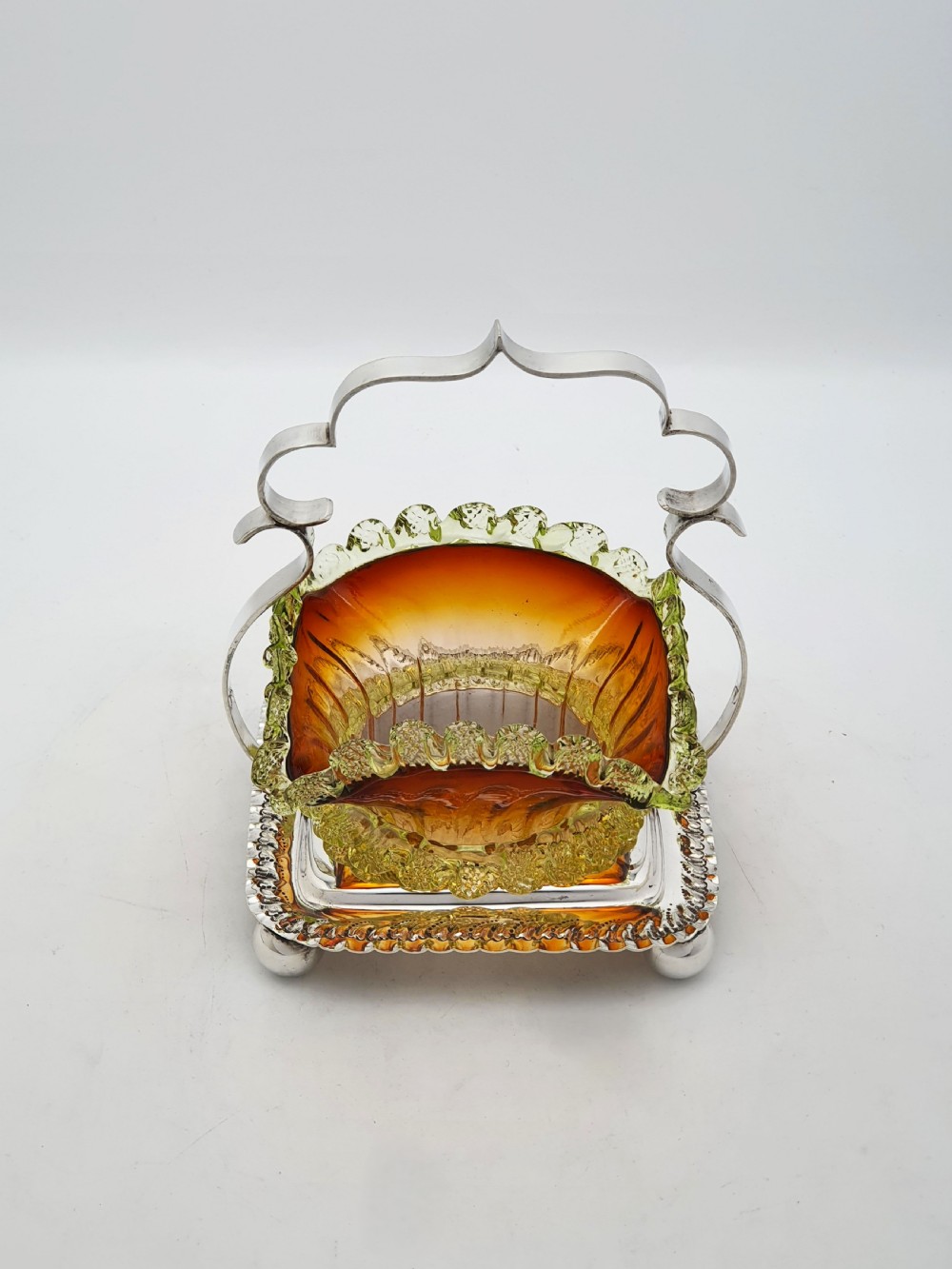 An Edwardian silver-plated stand with a shaped handle and ball feet holding a ruffled amber and yellow glass bonbon dish. View II