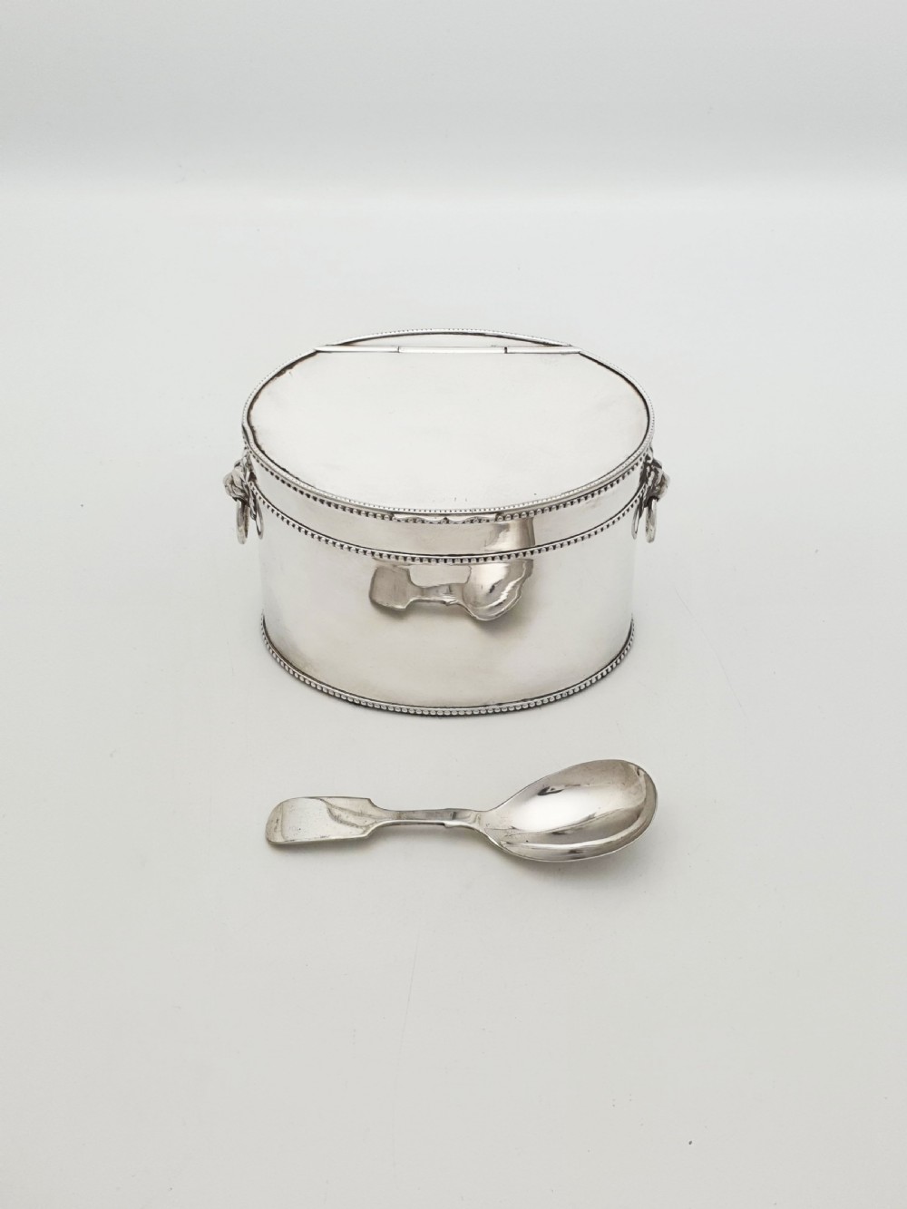 An oval silver tea caddy featuring lion mask ring handles on the sides, a hinged lid with beaded edging, and a front clip holding a small silver caddy spoon.