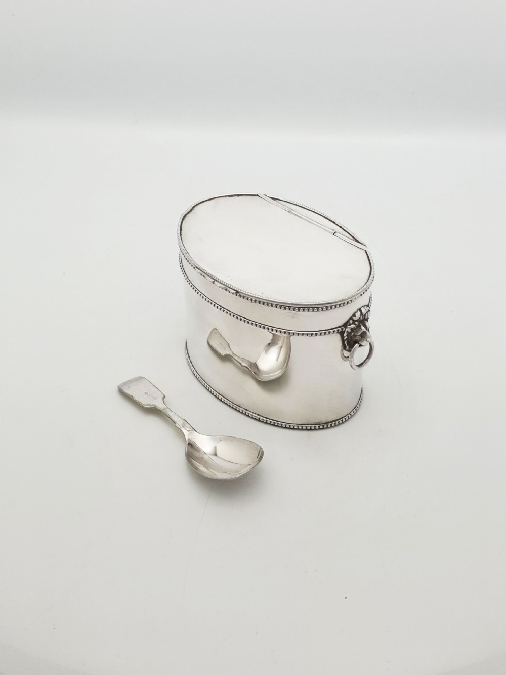 An oval silver tea caddy featuring lion mask ring handles on the sides, a hinged lid with beaded edging, and a front clip holding a small silver caddy spoon. View II