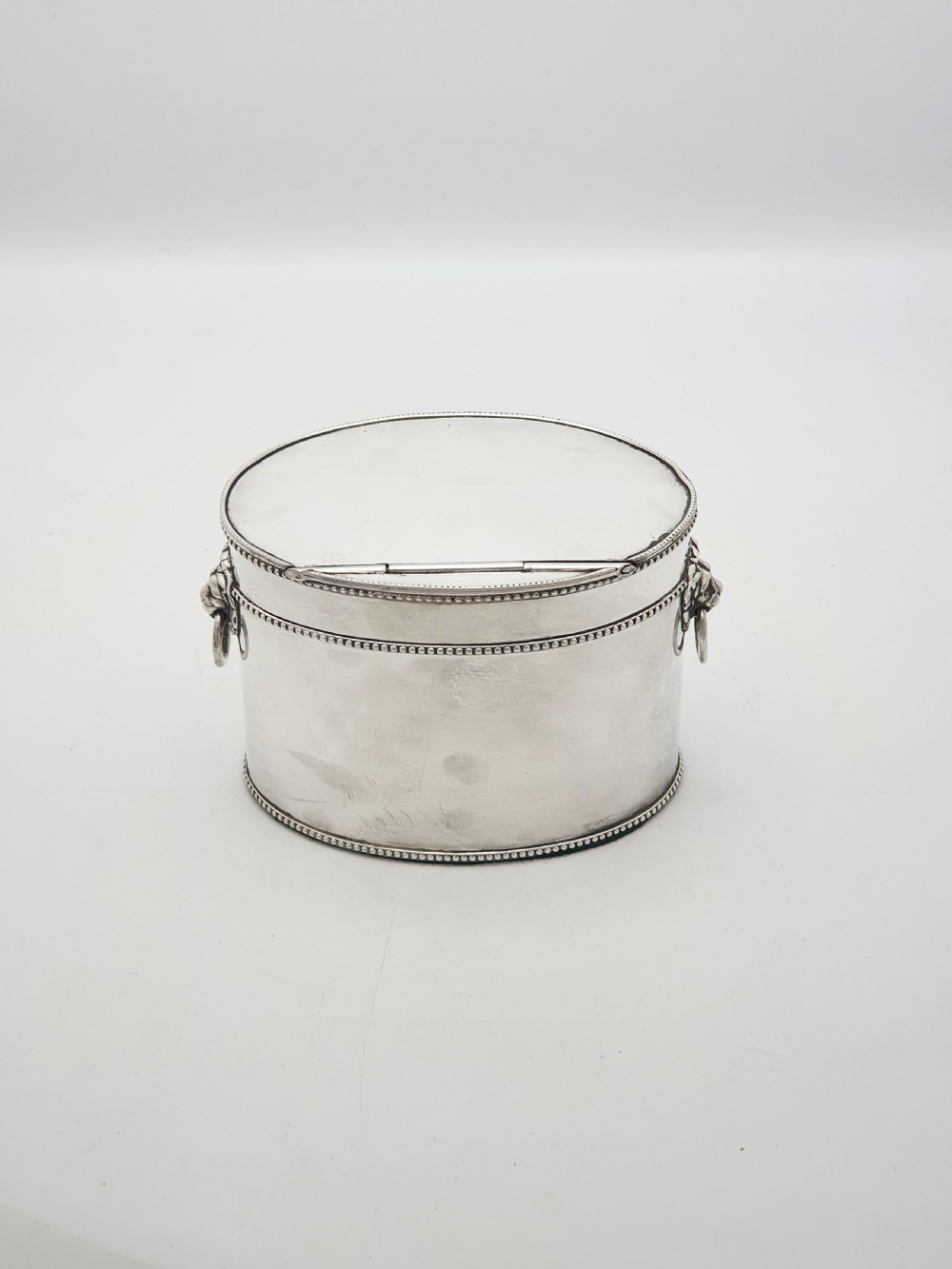 An oval silver tea caddy featuring lion mask ring handles on the sides, a hinged lid with beaded edging, and a front clip holding a small silver caddy spoon. View VII
