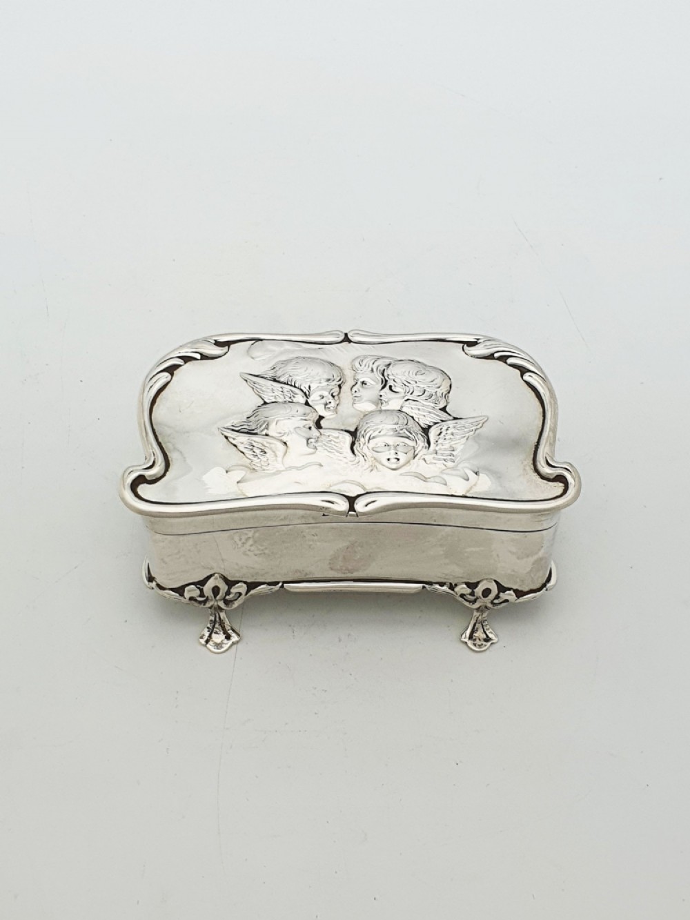 An Edwardian sterling silver trinket box featuring an embossed lid depicting cherub heads and a scrolling border, supported by four cast feet.