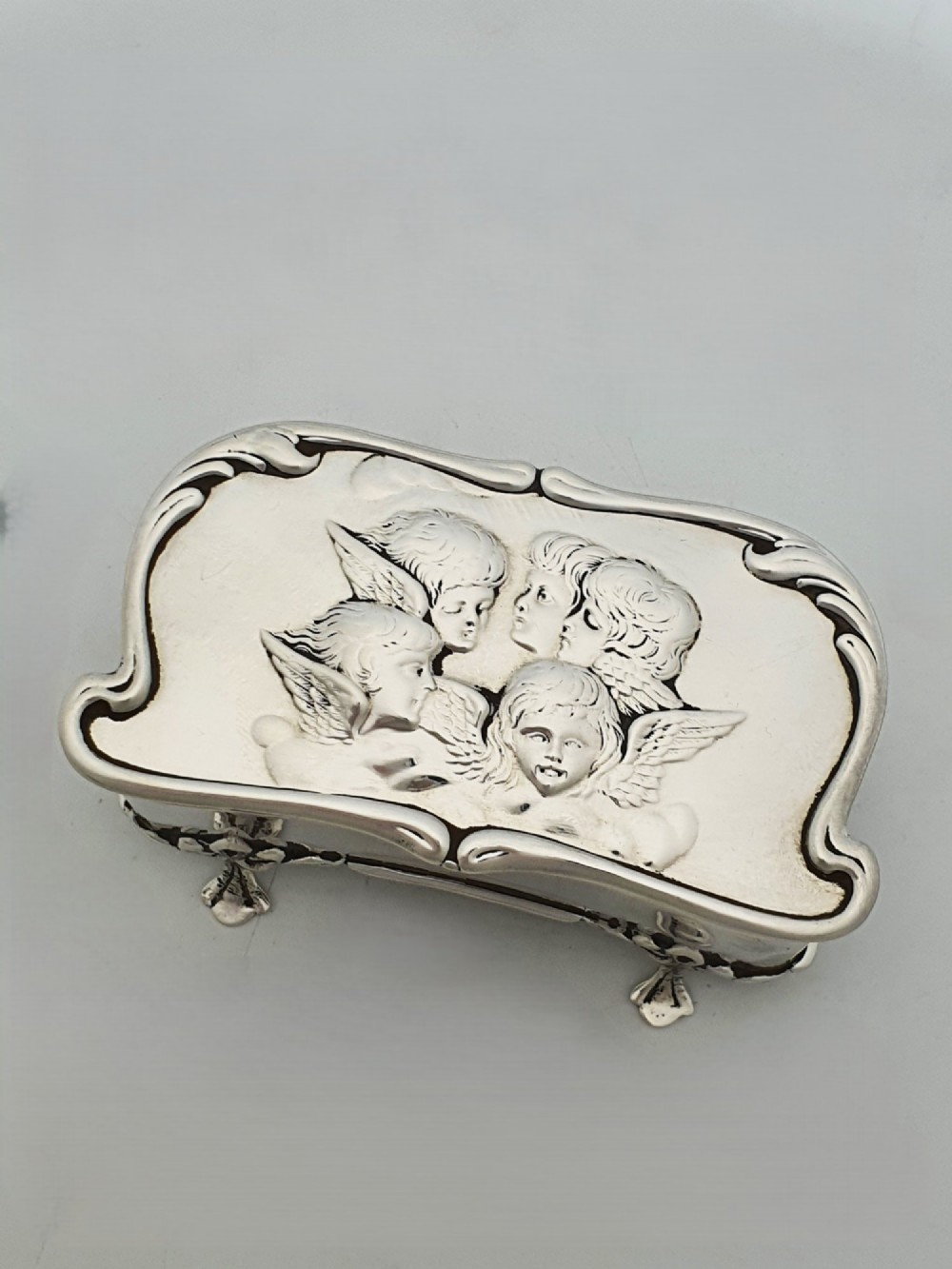 An Edwardian sterling silver trinket box featuring an embossed lid depicting cherub heads and a scrolling border, supported by four cast feet. View II