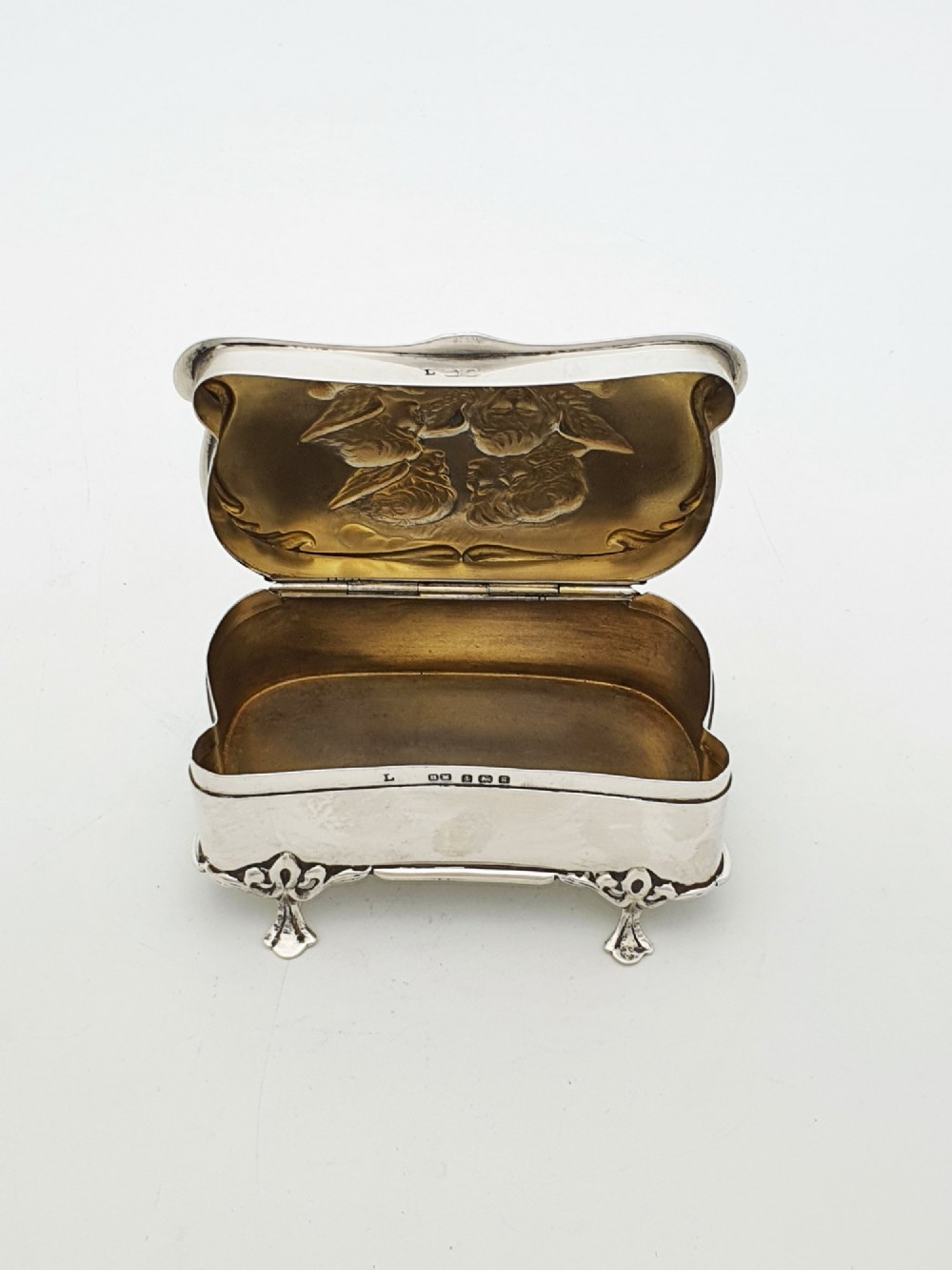 An Edwardian sterling silver trinket box featuring an embossed lid depicting cherub heads and a scrolling border, supported by four cast feet. View III