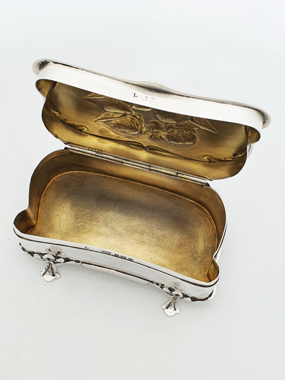 An Edwardian sterling silver trinket box featuring an embossed lid depicting cherub heads and a scrolling border, supported by four cast feet. View IV