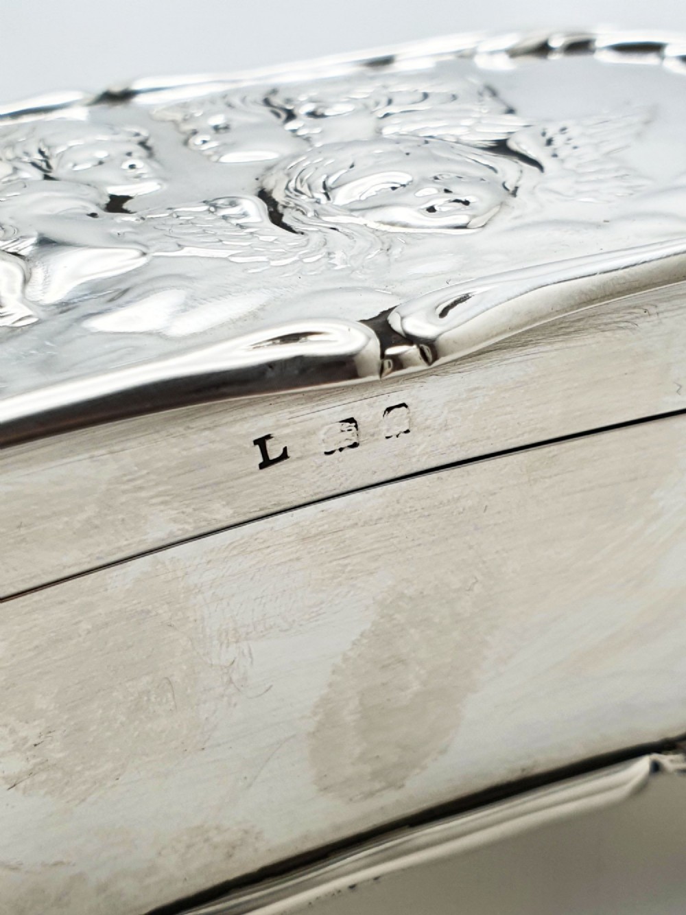 An Edwardian sterling silver trinket box featuring an embossed lid depicting cherub heads and a scrolling border, supported by four cast feet. View VI