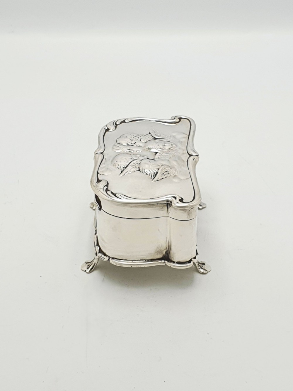 An Edwardian sterling silver trinket box featuring an embossed lid depicting cherub heads and a scrolling border, supported by four cast feet. View VII