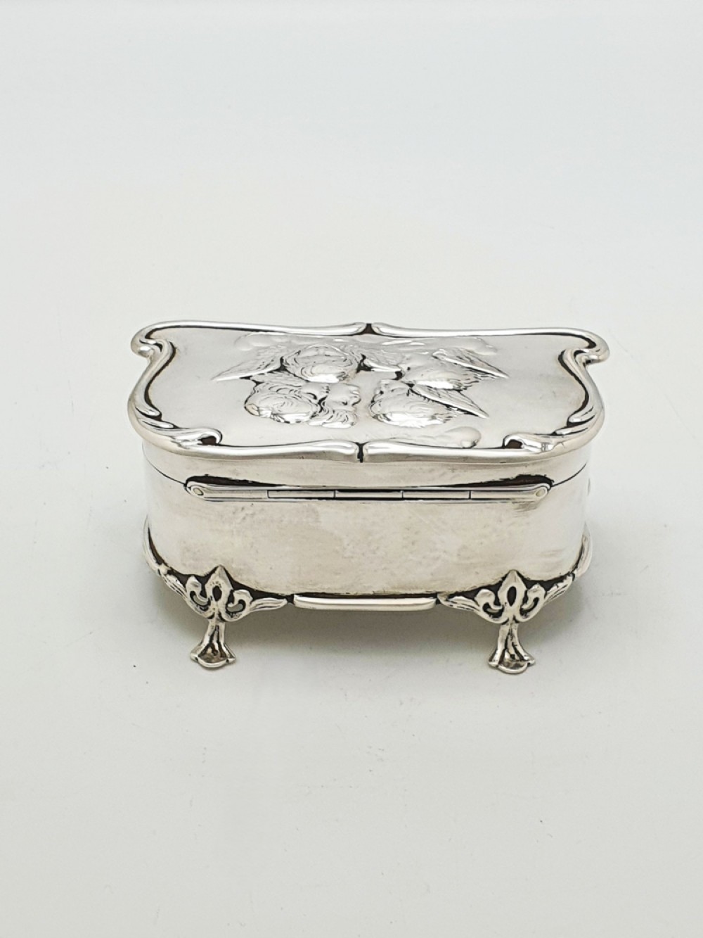 An Edwardian sterling silver trinket box featuring an embossed lid depicting cherub heads and a scrolling border, supported by four cast feet. View VIII