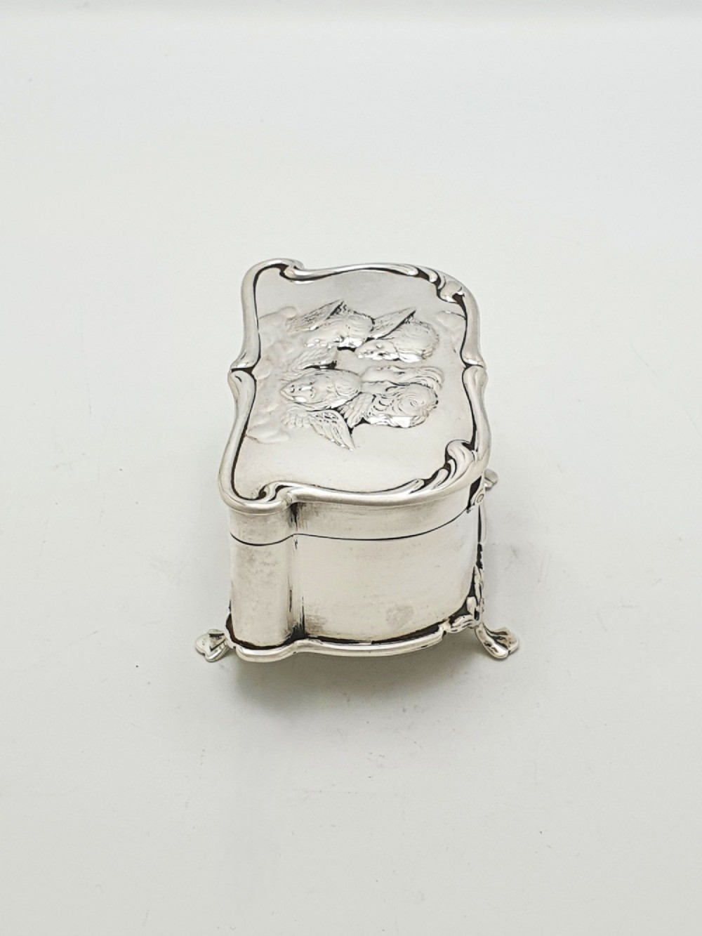 An Edwardian sterling silver trinket box featuring an embossed lid depicting cherub heads and a scrolling border, supported by four cast feet. View IX