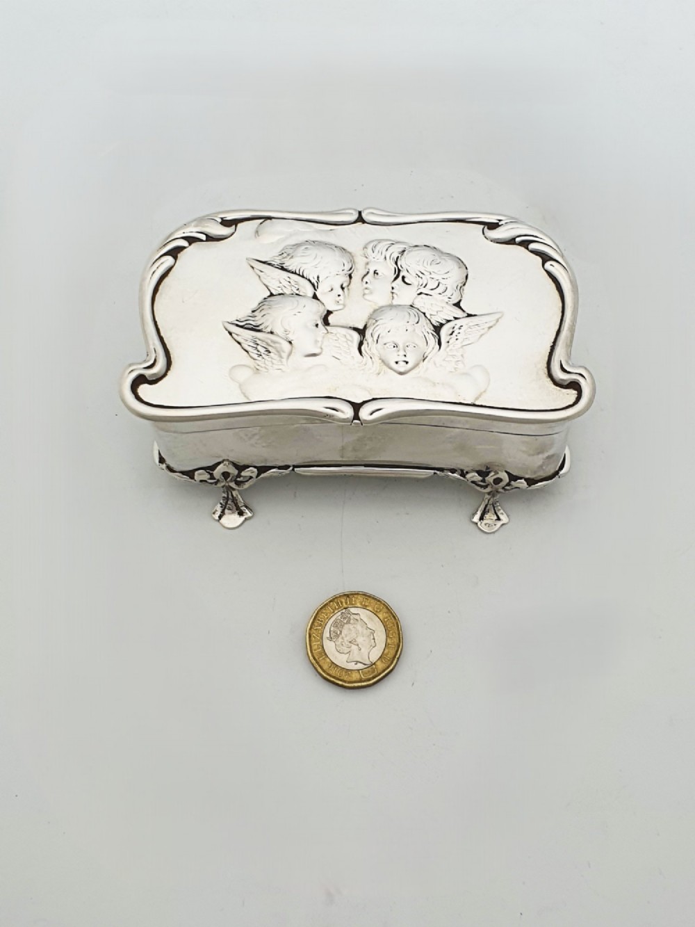 An Edwardian sterling silver trinket box featuring an embossed lid depicting cherub heads and a scrolling border, supported by four cast feet. View X