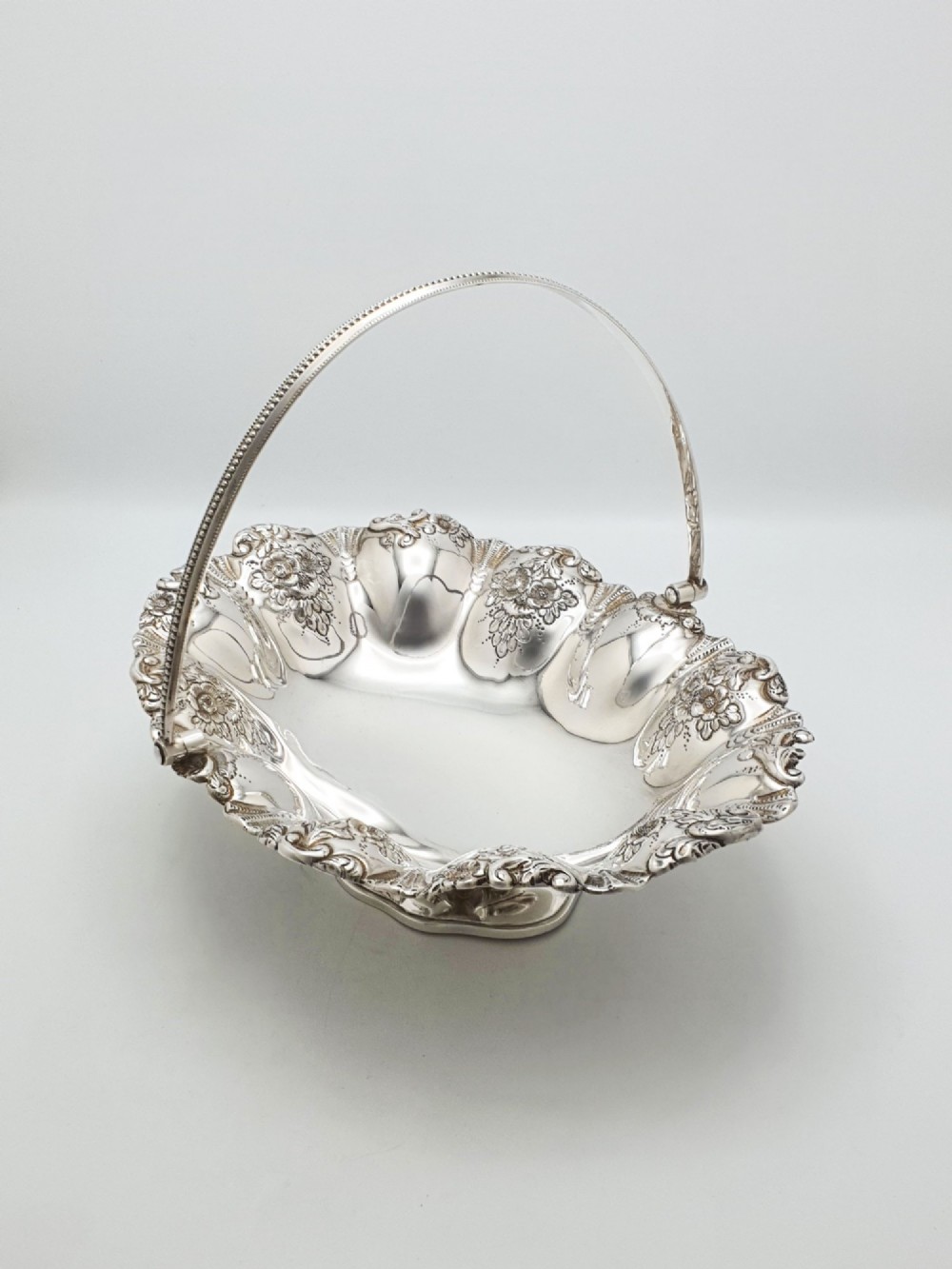 An Edwardian silver-plated oval basket featuring an overhead swing handle, fluted lobes, and a floral repousse rim.
