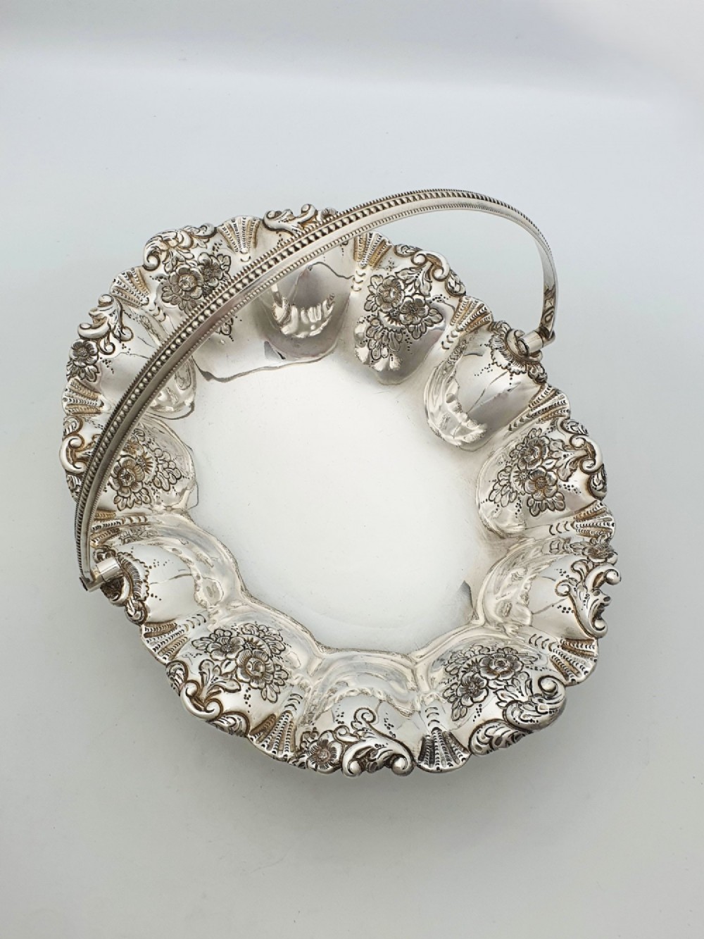 An Edwardian silver-plated oval basket featuring an overhead swing handle, fluted lobes, and a floral repousse rim. View II