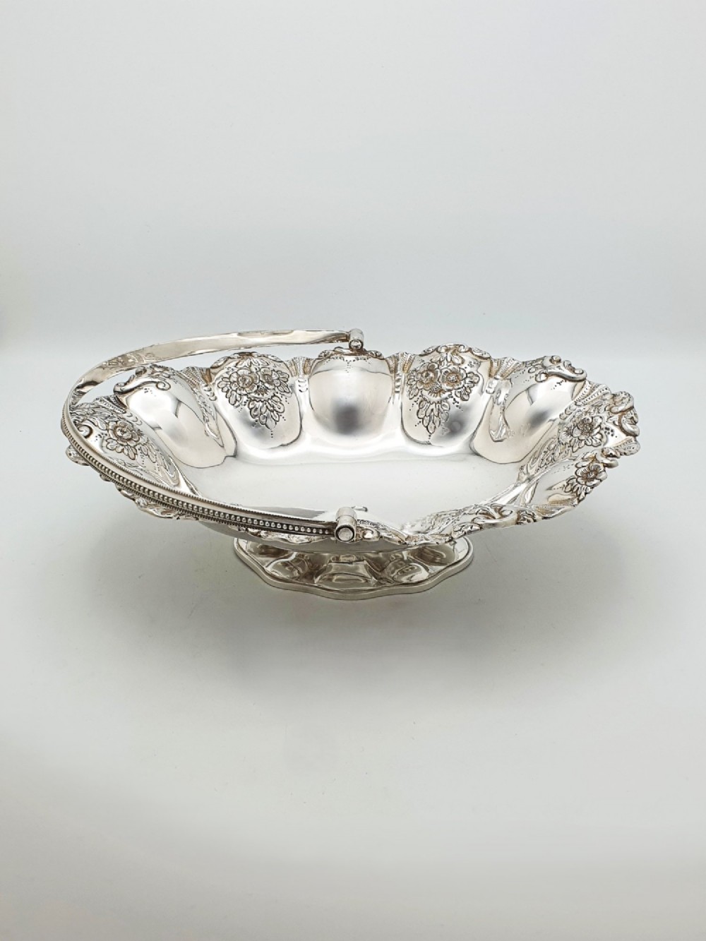An Edwardian silver-plated oval basket featuring an overhead swing handle, fluted lobes, and a floral repousse rim. View VI